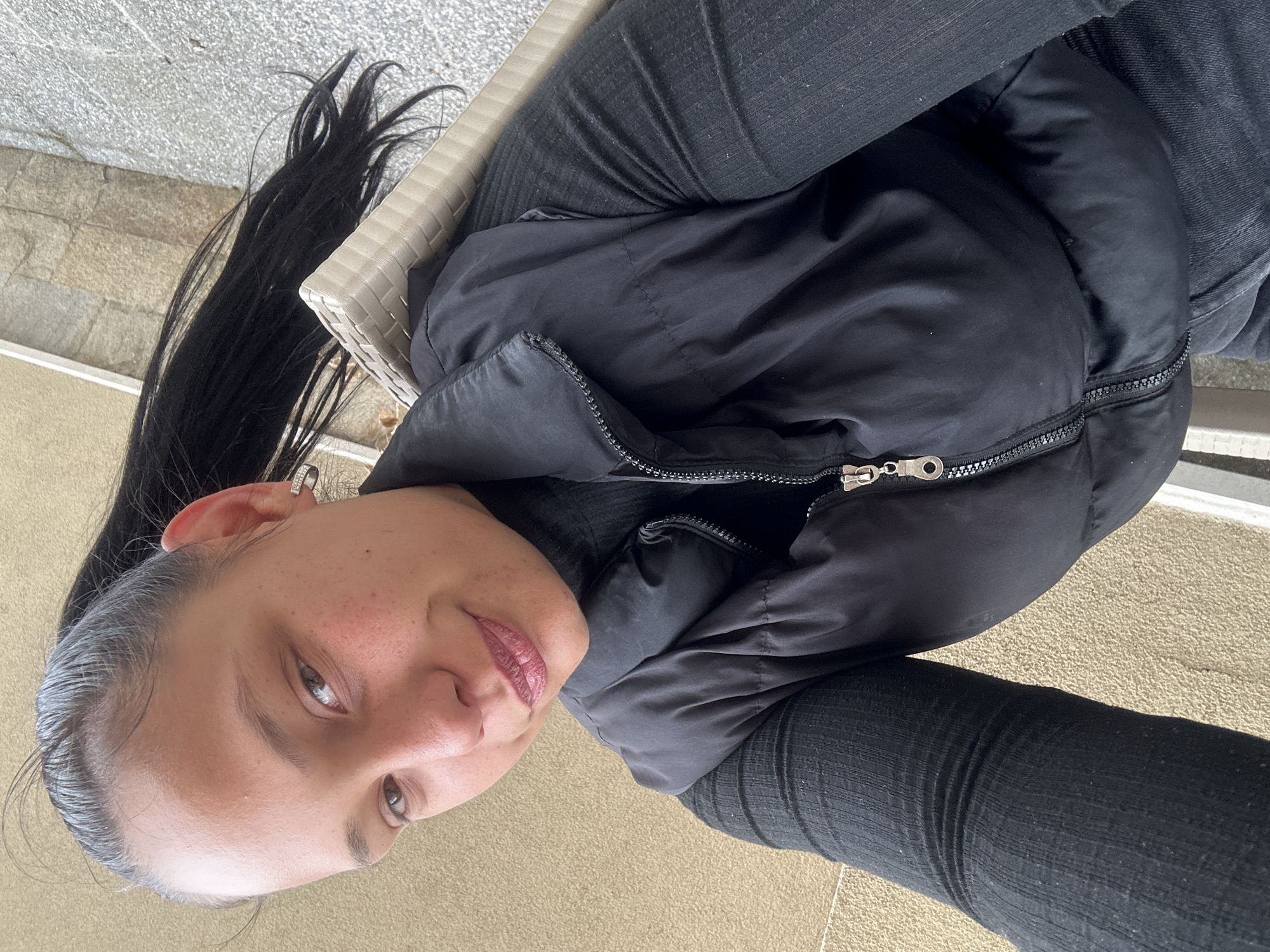 This media file features a portrait of a young person, likely female, captured from an elevated, oblique perspective. The subject is seated outdoors or in a semi-outdoor setting in Sofia, Bulgaria, looking towards the camera with a calm expression and a subtle smile.
She has dark, long hair, pulled back into a ponytail or bun, with a few loose strands visible. Her eyes are light brown, and she wears a pinkish-mauve lip color. Small freckles are noticeable on her face. A silver-colored hoop earring adorns her left ear.
Her attire consists of a black puffer jacket with a silver zipper and pull, a dark long-sleeved top worn underneath, and dark pants, possibly jeans or leggings. She is seated in a light-colored chair, parts of which are visible on the left side of the frame, showing a woven or wicker-like texture.
The background includes a light gray, textured surface, possibly a stone wall or paving, and a light beige/tan textured ground directly beneath her. The natural lighting indicates it is daytime. There is no visible text in the image.