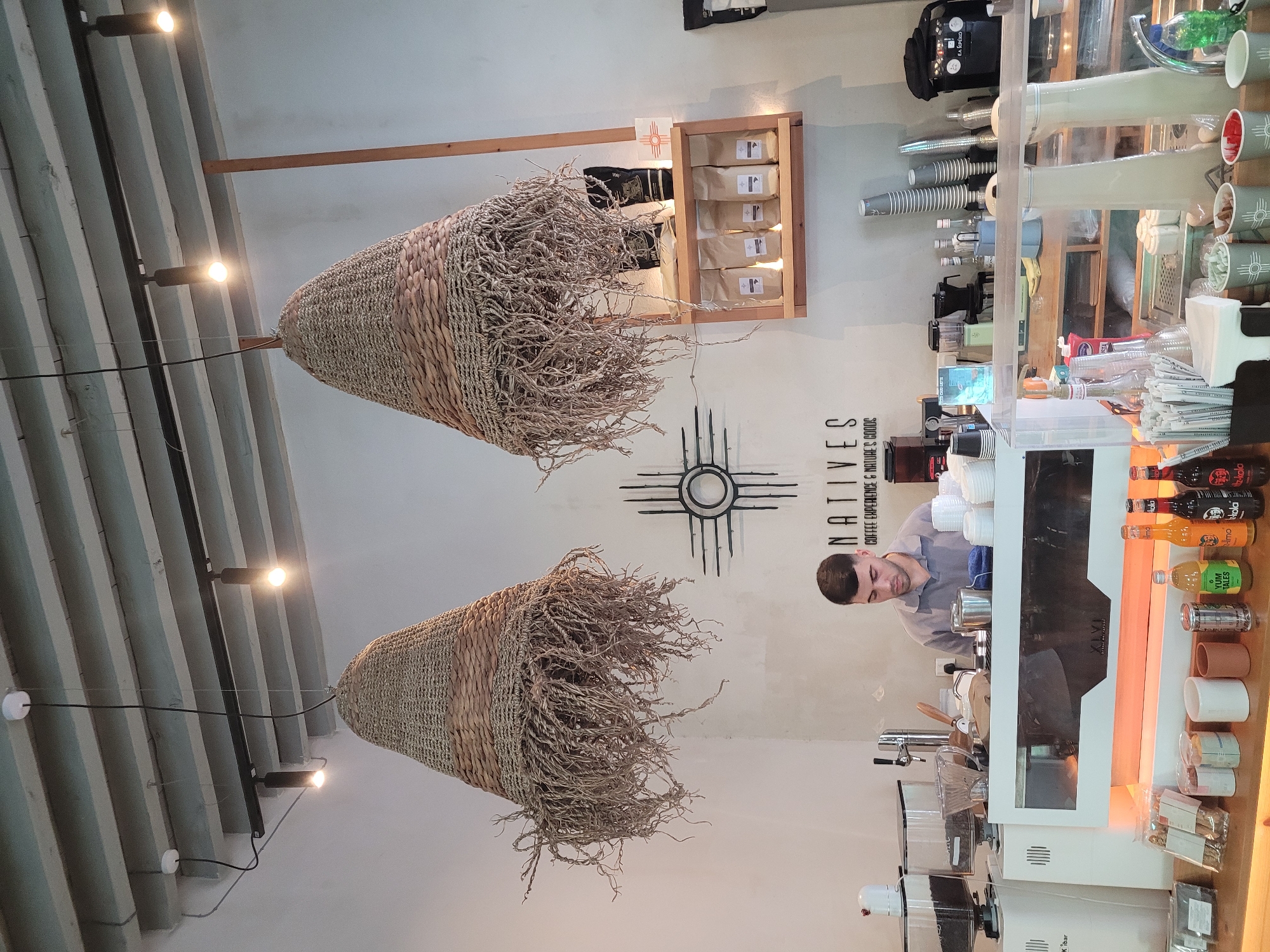 The image depicts the interior of a coffee shop named "NATIVES Coffee Experience & Nature's Goods" located in Limassol, Cyprus. In the center of the frame, a barista, a young man with short dark hair, is standing behind a counter, focused on his work. The coffee shop features a rustic ambiance with two large, woven lamps hanging from the ceiling. A distinctive sun symbol with four points is mounted on the white wall behind the counter. Shelves on the right side of the wall display bags of coffee. The counter area is equipped with various coffee-making appliances, including grinders and espresso machines. On the wooden counter in the foreground, there are bottles of beverages, cups, and other coffee shop essentials. The overall scene suggests a modern, well-lit coffee establishment with a focus on natural elements.