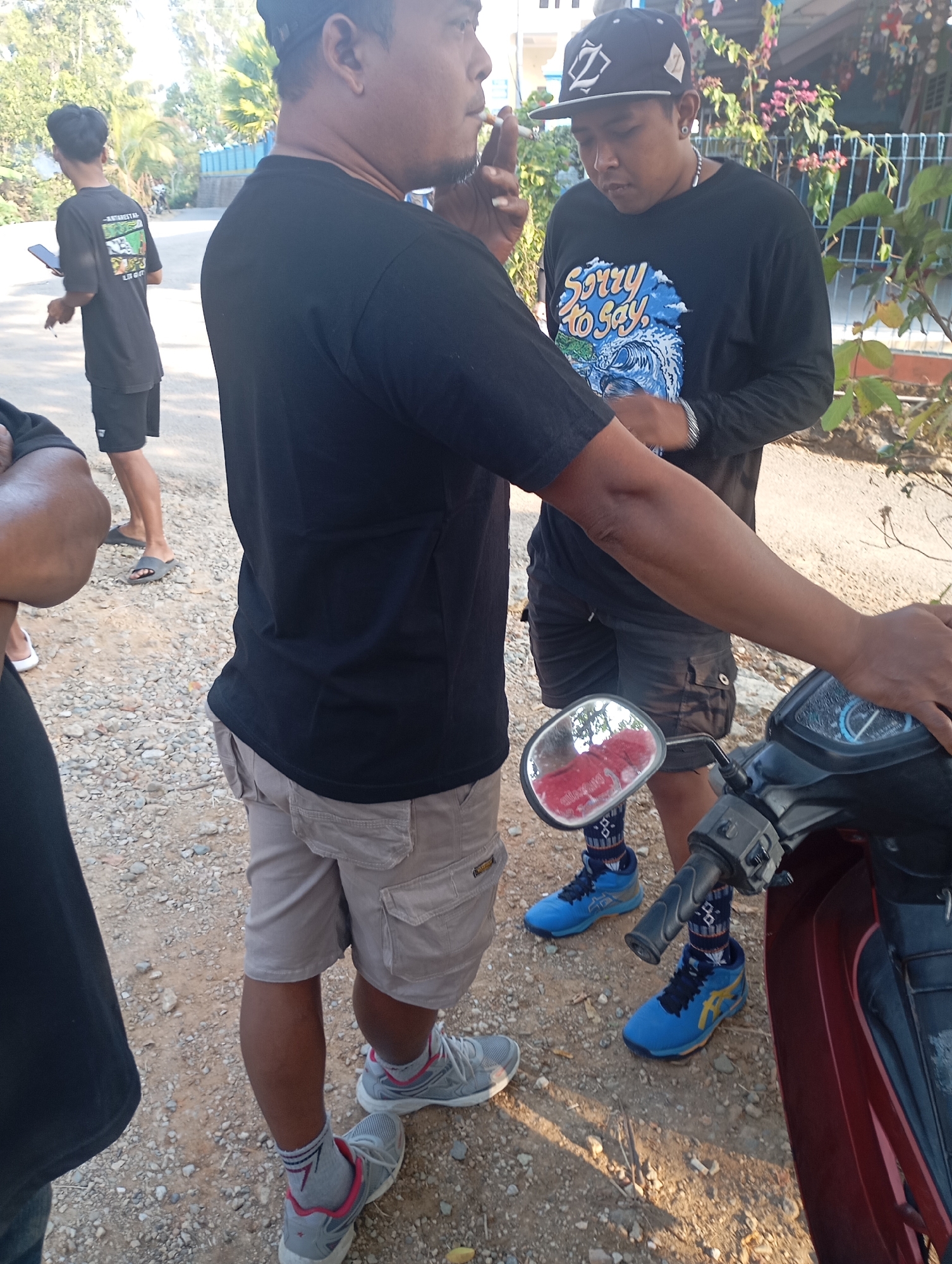 The image depicts a group of people standing on a gravel-covered surface, likely a roadside in Ketepung, Indonesia. In the foreground, a person wearing a black t-shirt and light-colored cargo shorts is partially visible on the left. To their right, another individual in a black long-sleeved shirt with the text "Sorry to Say" and a graphic print is standing, looking down and holding a cigarette. This person is also wearing a black baseball cap. A motorcycle, primarily its handlebars and part of its body, is visible on the right side of the frame, with a red reflection visible in its side mirror. In the background, another person in a black t-shirt and shorts is seen walking away from the camera. The environment appears to be outdoors, with foliage and a building visible in the distance. The lighting suggests it is daytime, with sunlight casting shadows.