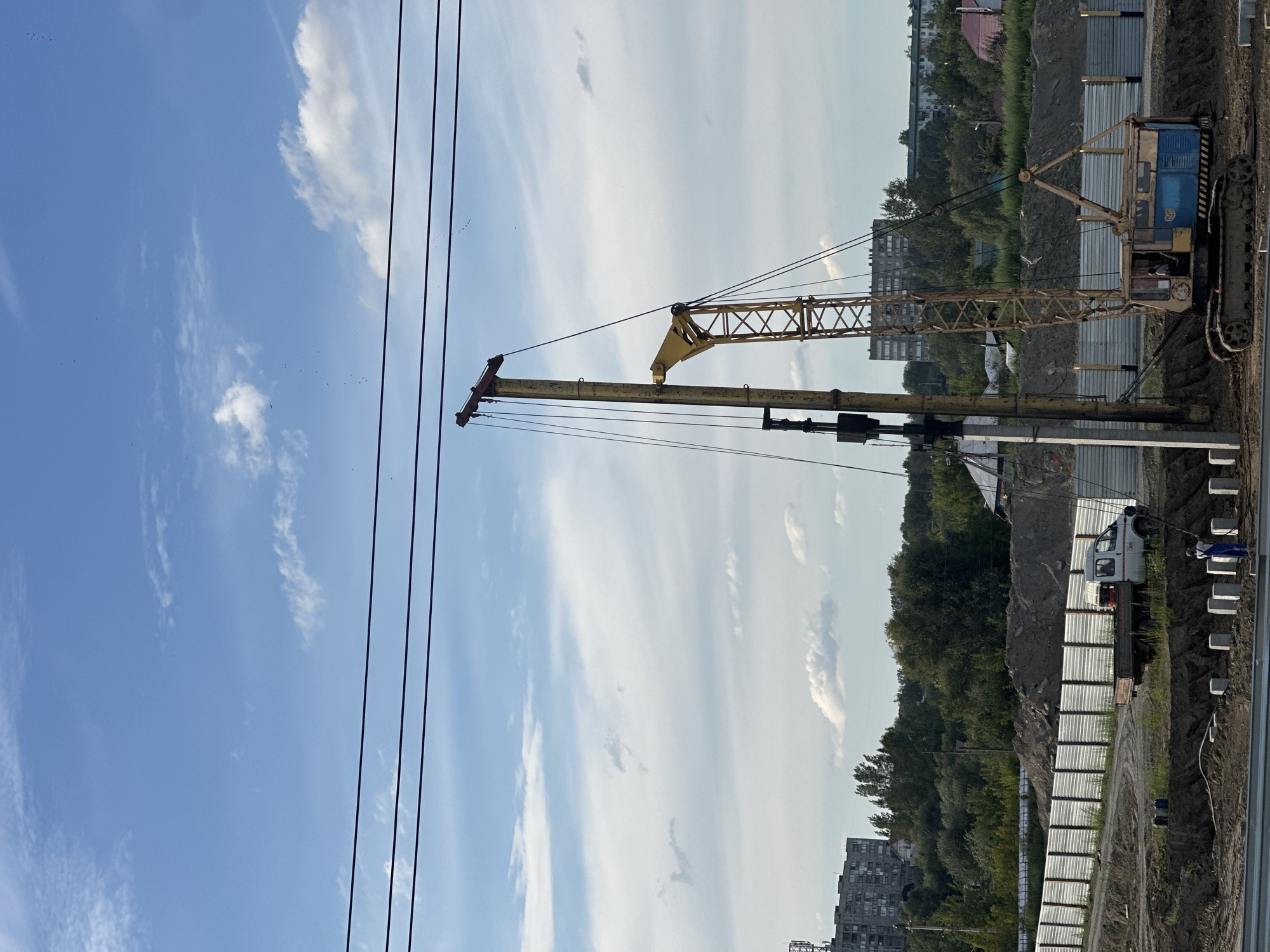 A construction site is depicted in Biysk, Russia, under a partly cloudy blue sky. In the foreground, a large, yellow crane with a blue cab sits on a tracked base. Next to it, a tall yellow piling rig with a concrete pillar positioned vertically is being operated. A white pickup truck is parked behind a corrugated metal fence, with a person in blue overalls visible in the foreground. In the background, several residential buildings and green trees are visible. Overhead, three dark power lines stretch across the frame.