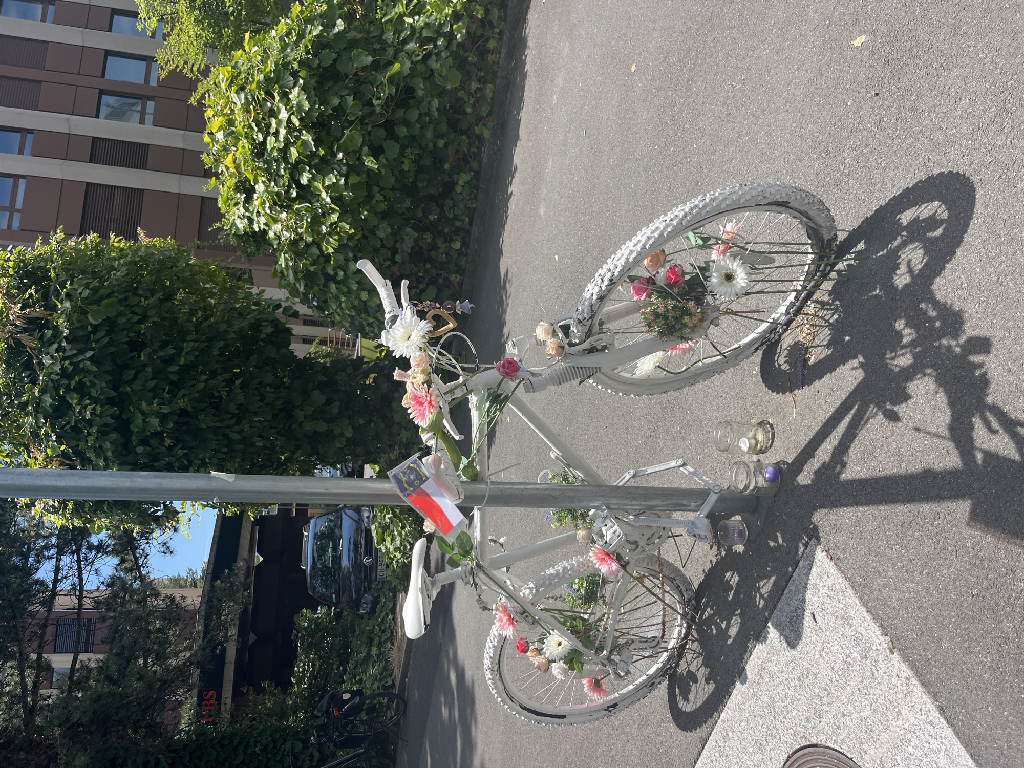 A white bicycle, adorned with numerous artificial flowers, is parked and attached to a metal pole in Geneva, Switzerland. The bicycle is decorated with pink, white, and red flowers, and a small flag with red and white stripes is attached to the handlebars. Two glass jars, one containing a purple object and the other empty with a lit candle, are placed on the ground near the bicycle's front wheel. In the background, a dark-colored car is visible, along with a building with brown siding and windows. Lush green foliage surrounds the scene, suggesting an outdoor urban environment. The ground is paved with asphalt.