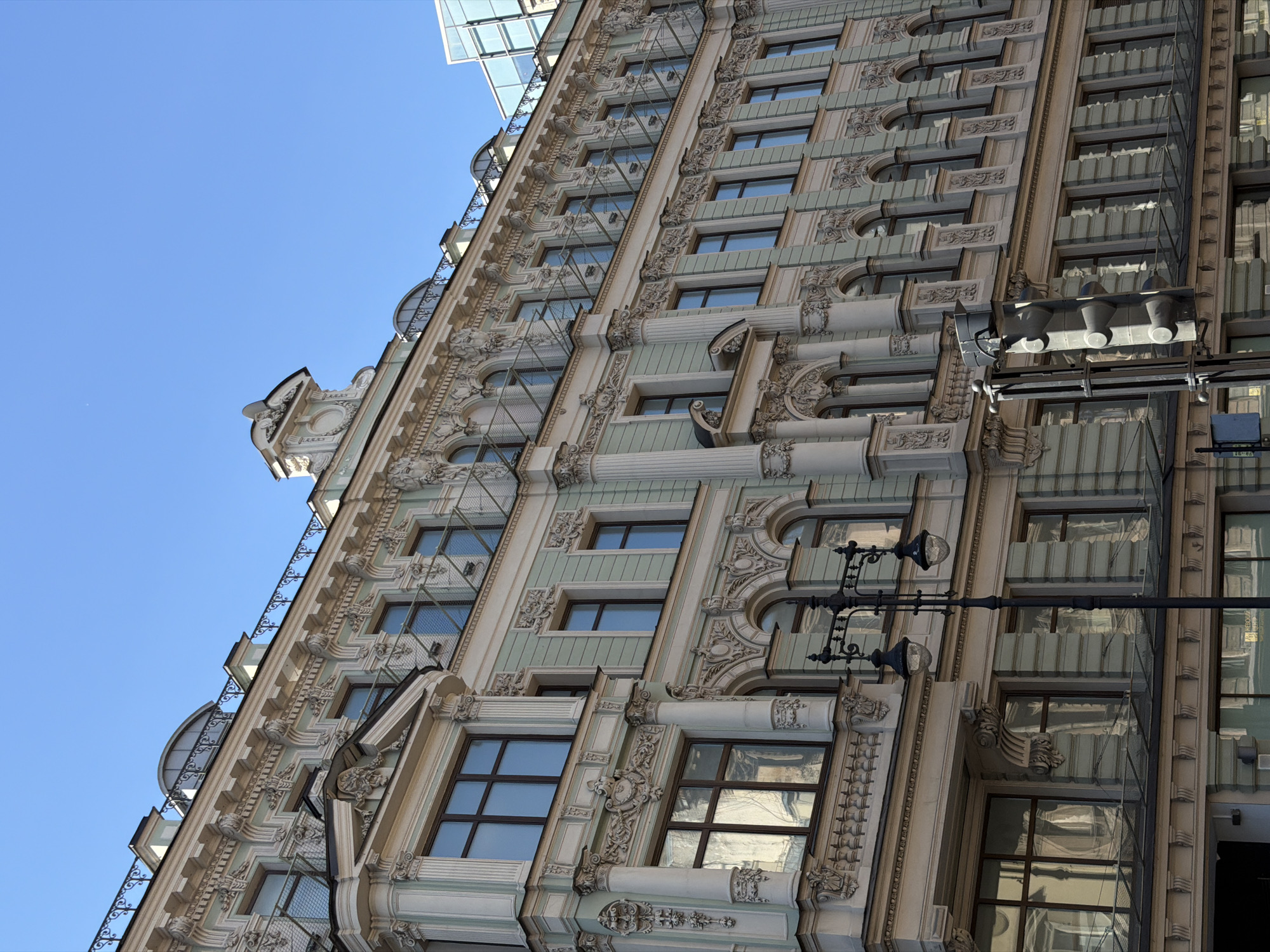 The image shows an ornate, light green and cream-colored building with multiple floors and numerous windows, exhibiting classical architectural details such as columns, intricate moldings, and decorative carvings. The building appears to be a historic structure. A black street lamp with two globes stands in the foreground, to the left of a traffic signal. The sky is clear and blue. The building is located in Saint Petersburg, Russia, on Nevsky Prospekt, a prominent street in the city. There are no people visible in the image. The scene is a street view of an urban environment.