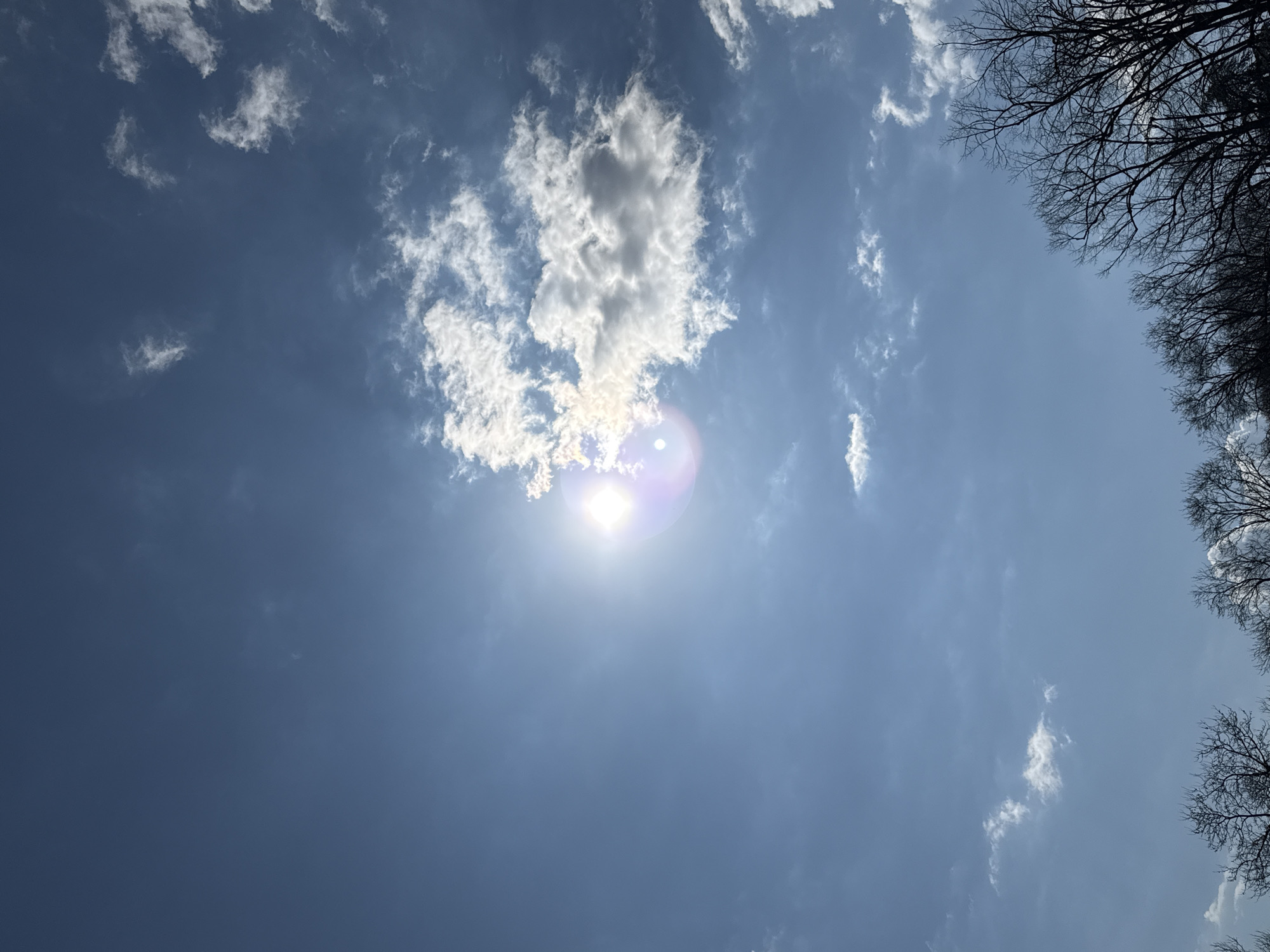 The image shows a bright, daytime sky with a few scattered cumulus clouds. The sun is visible, obscured by a thin cloud, creating a lens flare effect. Silhouetted tree branches are visible in the upper right corner of the image. The location is likely outside, as the focus is on the sky and natural elements. The specific location within Varna, Bulgaria cannot be determined from this image alone. There are no people visible in the image.