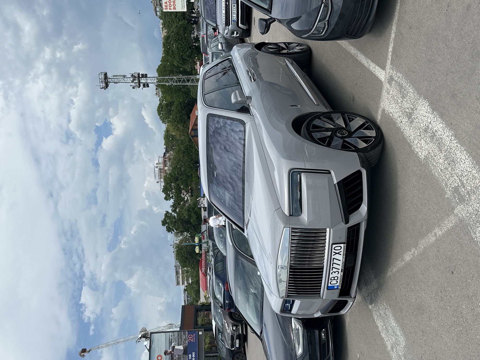 The image captures a parked Rolls-Royce Cullinan, with the license plate "CB 3777 XO," amidst other vehicles in a parking area. The scene includes additional cars like a Range Rover, and an Audi. In the background, the cityscape features buildings and greenery. A tall structure with a boom and gondola, possibly an amusement park ride or observation tower, is visible against a cloudy sky.
The license plate indicates the car is registered in Bulgaria.
The license plate of the Range Rover is "CT 01 CCM".
There is also a sign for "SEA FOOD BOIL" and a billboard advertising "2C technology".
The number on the billboard is "82691HP".
There is a man on the billboard wearing a white shirt.
Based on the Cyrillic license plate, the billboard, and the general appearance, the photograph was most likely taken in the city of Varna, Bulgaria.