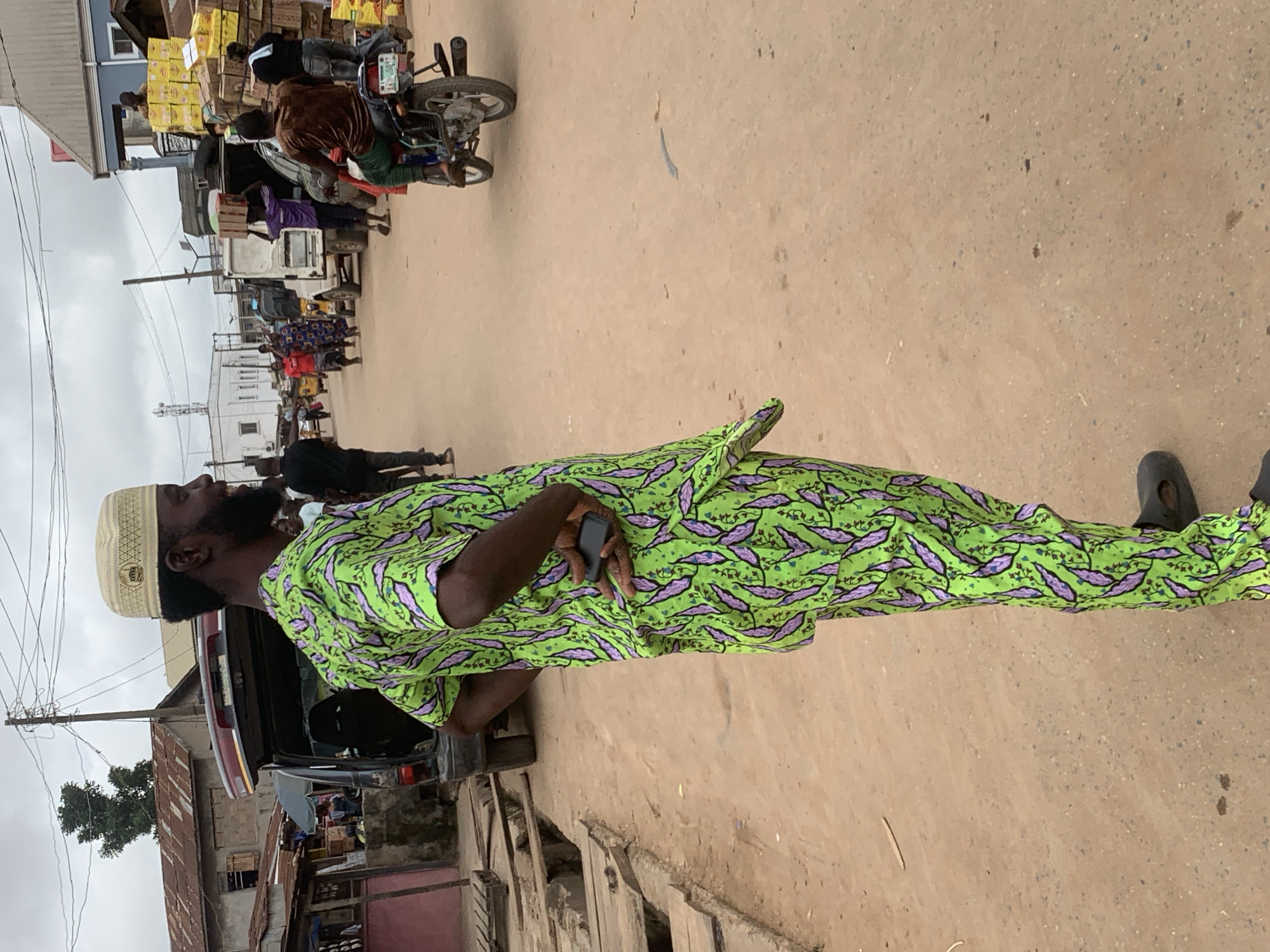 The image depicts a street scene in Shagamu, Nigeria, likely a market area. The foreground is dominated by a man facing away from the camera, wearing a brightly colored green and purple patterned traditional Nigerian outfit and a light-colored cap. He is holding a dark-colored mobile phone in his right hand, tucked behind his back.
The environment is a dusty, unpaved street with visible drainage ditches lined with wooden planks on the left side. Buildings with corrugated iron roofs and some concrete structures are visible in the background. Several other people are present, including individuals walking, carrying goods, and a person riding a motorcycle. Vehicles, including an open trunk van and what appears to be a white delivery truck, are also in the scene. Stacks of yellow boxes, possibly merchandise, are piled high near a blue building on the right side of the street. The sky is overcast.