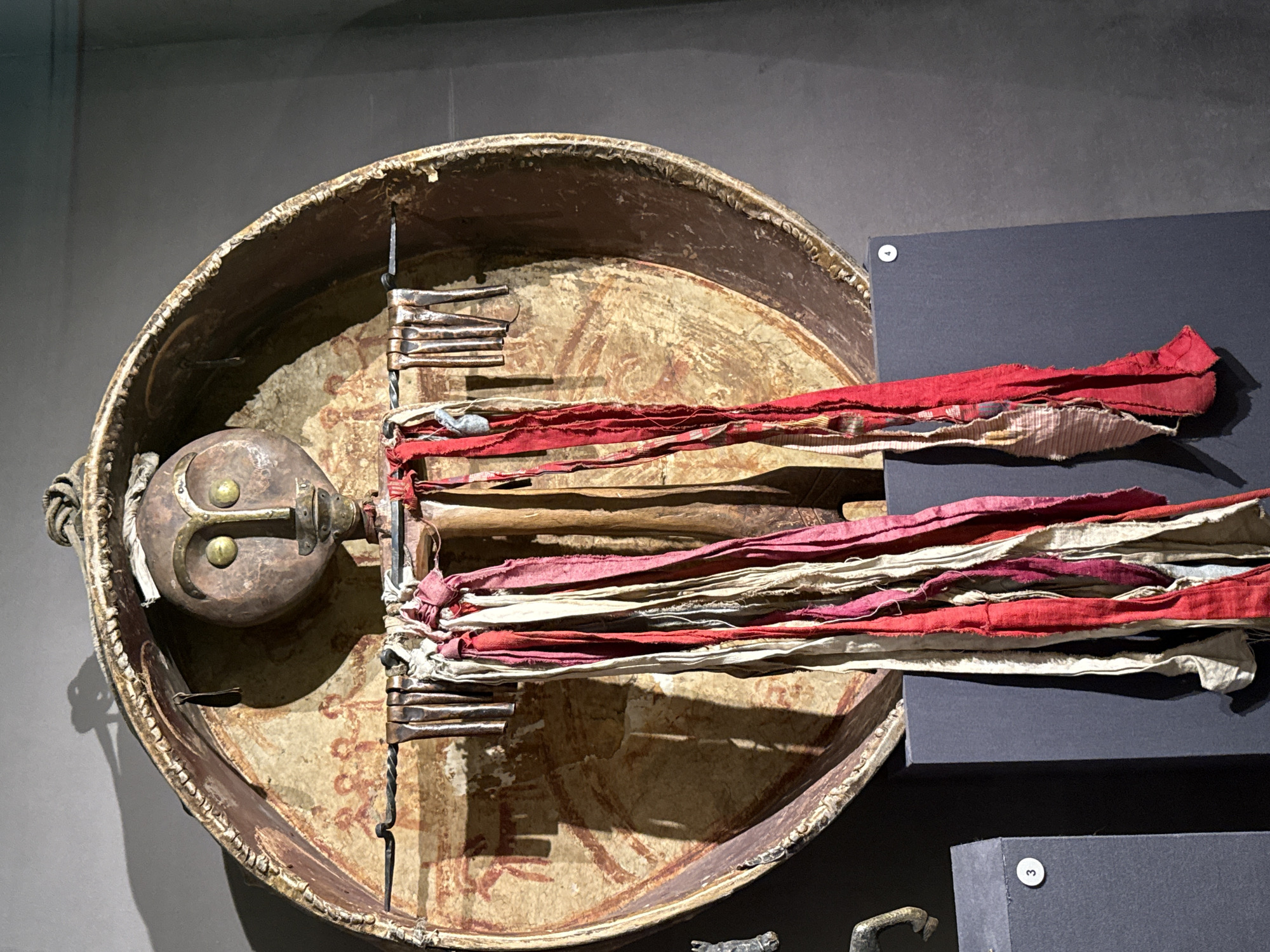 This image shows a ritualistic object, likely a mask or effigy, displayed in a museum setting. The object features a central, stylized face with large, round, yellow eyes and a prominent nose and mouth. The face is mounted on a large, circular, wooden frame with painted designs on its interior surface. Attached to the front of the frame are numerous strips of fabric in various shades of red, pink, and off-white, hanging down like a skirt. The object is suspended against a dark gray wall, with subtle lighting casting shadows. The background also shows a portion of a dark, rectangular display pedestal, with a circular label marked with the number "4".
While the image itself does not explicitly state its location, it is important to note that this object is displayed at the Kunstkamera in Saint Petersburg, Russia. The Kunstkamera is Russia's first public museum, founded by Peter the Great in 1714, and it houses a vast collection of ethnographic and anthropological artifacts from around the world. The object in the image is characteristic of the indigenous art and cultural practices collected by the museum.