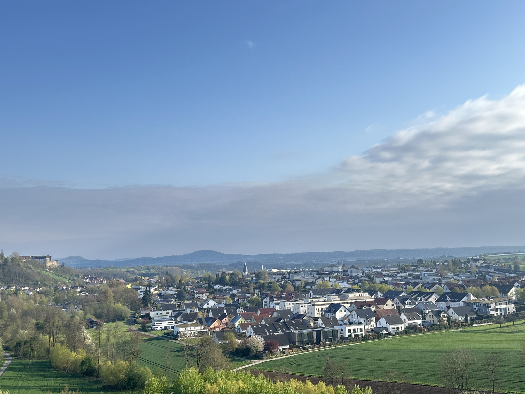 The image shows a panoramic view of a town nestled within a landscape of rolling hills and open fields. The foreground features vibrant green fields and trees in various stages of spring foliage. The town itself is characterized by a mix of residential buildings, with varying roof colors and architectural styles. A church steeple rises prominently from the town's center. In the distance, a castle-like structure is perched atop a hill, overlooking the scene. The sky is a gradient of blue, with some scattered clouds obscuring the horizon. The scene is bathed in soft, natural light. The location is in the city of Unknown, Unknown.