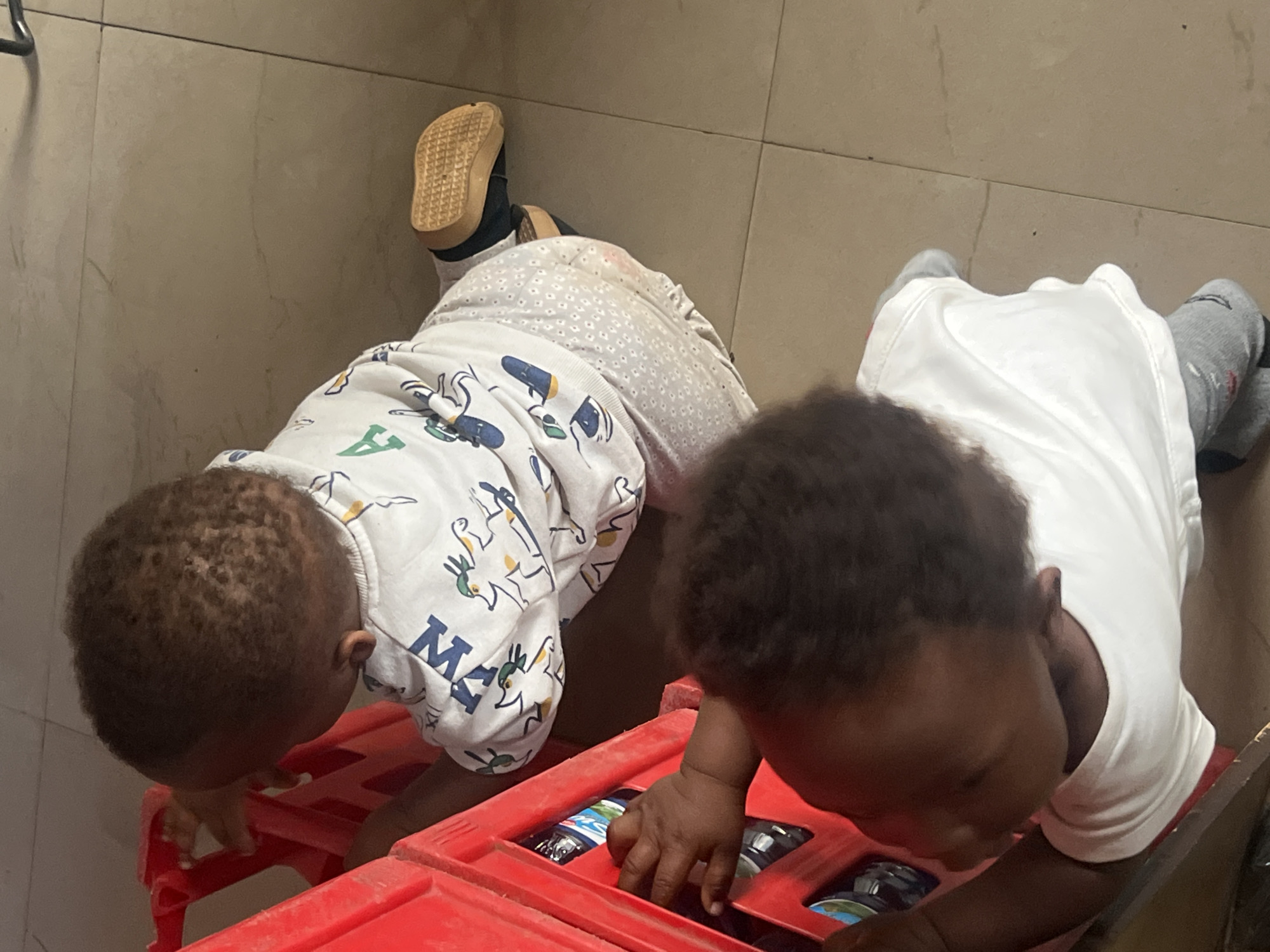 Two young children are on the floor. One is wearing white patterned pajamas and one shoe, and the other is wearing a white top and grey socks. They are both looking down into a red plastic crate. The location is Gura Top, Nigeria. The specific details or setting within Gura Top are not distinguishable from the image.