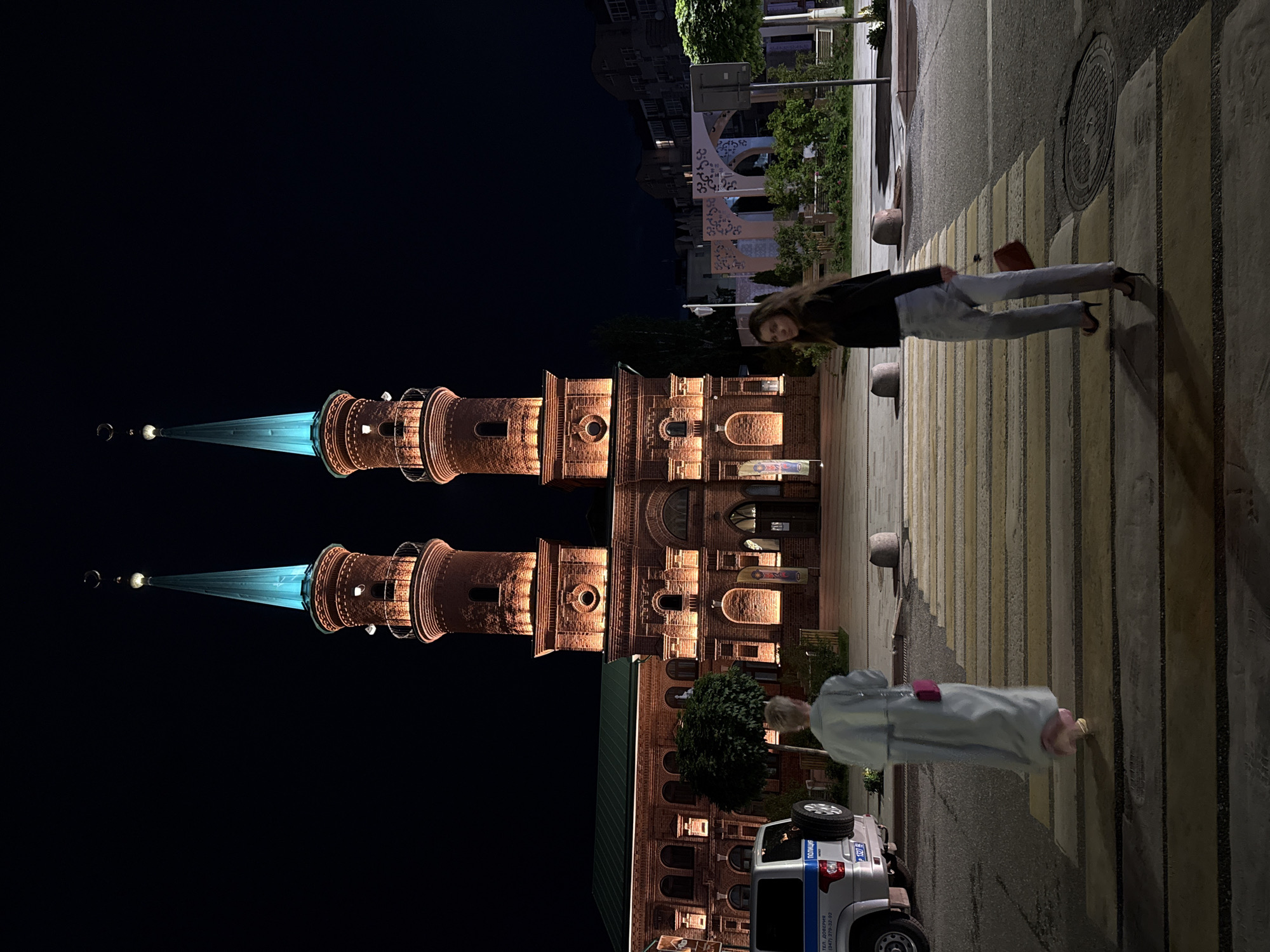 The photograph captures a nighttime scene outside a mosque in Ufa, Russia, likely the Ufa Cathedral Mosque, judging by its distinct architecture featuring two illuminated minarets topped with crescent moons. The mosque's brick facade is warmly lit, highlighting its intricate details and arched entrances. In the foreground, a pedestrian crossing is visible, marked by yellow and white stripes.
Two women are present in the scene. The first, closer to the left of the frame, is seen from behind, walking away from the viewer and towards the mosque. She is wearing a long, light-colored trench coat and carries a small, pink handbag. The second woman is positioned in the right half of the frame, also walking across the pedestrian crossing, but facing more towards the viewer. She is wearing a dark top, light-wash jeans, and high heels, and carries a red handbag.
To the left of the pedestrian crossing, a silver police vehicle with visible markings, including "ПОЛИЦИЯ" (Police), is parked. The surrounding environment includes trees and some urban structures in the background, with streetlights illuminating parts of the scene. The overall atmosphere is one of urban nocturnal illumination, with the mosque serving as the central architectural element.