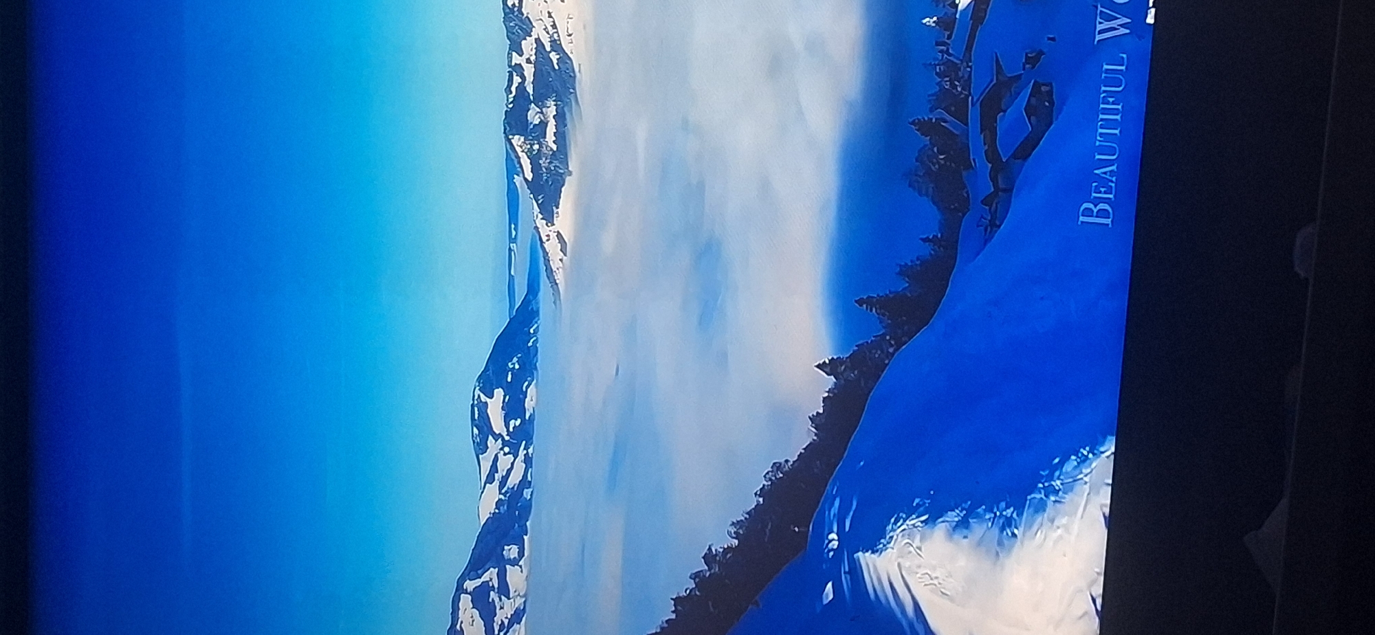 The image depicts a serene winter landscape with snow-covered mountains and a sea of clouds. In the foreground, a snowy slope leads down to a cluster of houses with snow-laden roofs, suggesting a small village nestled in the mountains. The sky is a clear, vibrant blue, transitioning to a lighter hue towards the horizon. The text "BEAUTIFUL W" is visible in white, serif font at the bottom right of the image, indicating it might be from a video or a title card. The scene is devoid of people. The specific location in Bila Tserkva, Ukraine is not identifiable from the image, as Bila Tserkva is not known for such mountainous, cloud-filled winter landscapes.