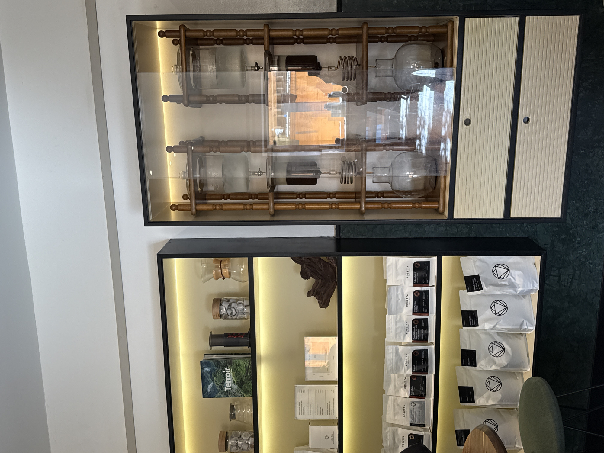 The scene depicts an interior view of a specialty coffee establishment, specifically identified by the "ROSETTA" branding on coffee bags, located in Cape Town, South Africa. The image is dominated by two distinct wall-mounted display units against a dark green wall and a white ceiling.
On the left, a black-framed shelving unit with three illuminated shelves displays various coffee-related items. The top shelf holds glass jars with wooden lids containing coffee pods, a clear glass jar, a book titled "Terroir" with a mountain landscape cover, an AeroPress coffee maker, and a clear glass Chemex-style brewer. The middle shelf features white menu cards or information sheets, a small white box, and a piece of dark, gnarled wood. The bottom shelf displays two rows of white pouches, each branded "ROSETTA" and labeled with coffee origins (e.g., "Chelele," "Ragati," "Benti Nenka," "Galende AA"). Some bags on the lower row also feature a geometric logo (a triangle within a circle). A dark green upholstered stool is partially visible in the lower left foreground.
On the right, a black-framed glass display cabinet houses two identical Hario-style cold brew coffee towers, each constructed with light brown wooden frames. Each tower features three levels: a top clear glass reservoir filled with water (one marked "3000 ml"), a middle glass chamber containing dark liquid (coffee concentrate) with a visible coiled metal component, and a bottom clear glass carafe. Below the glass section, two light-colored, horizontally ridged drawers with circular black pulls are visible. Reflections on the glass front of this cabinet include warm-toned architectural elements from the opposing side of the room.
No individuals are present in the image. The environment is clean, well-lit, and designed for coffee retail and display.