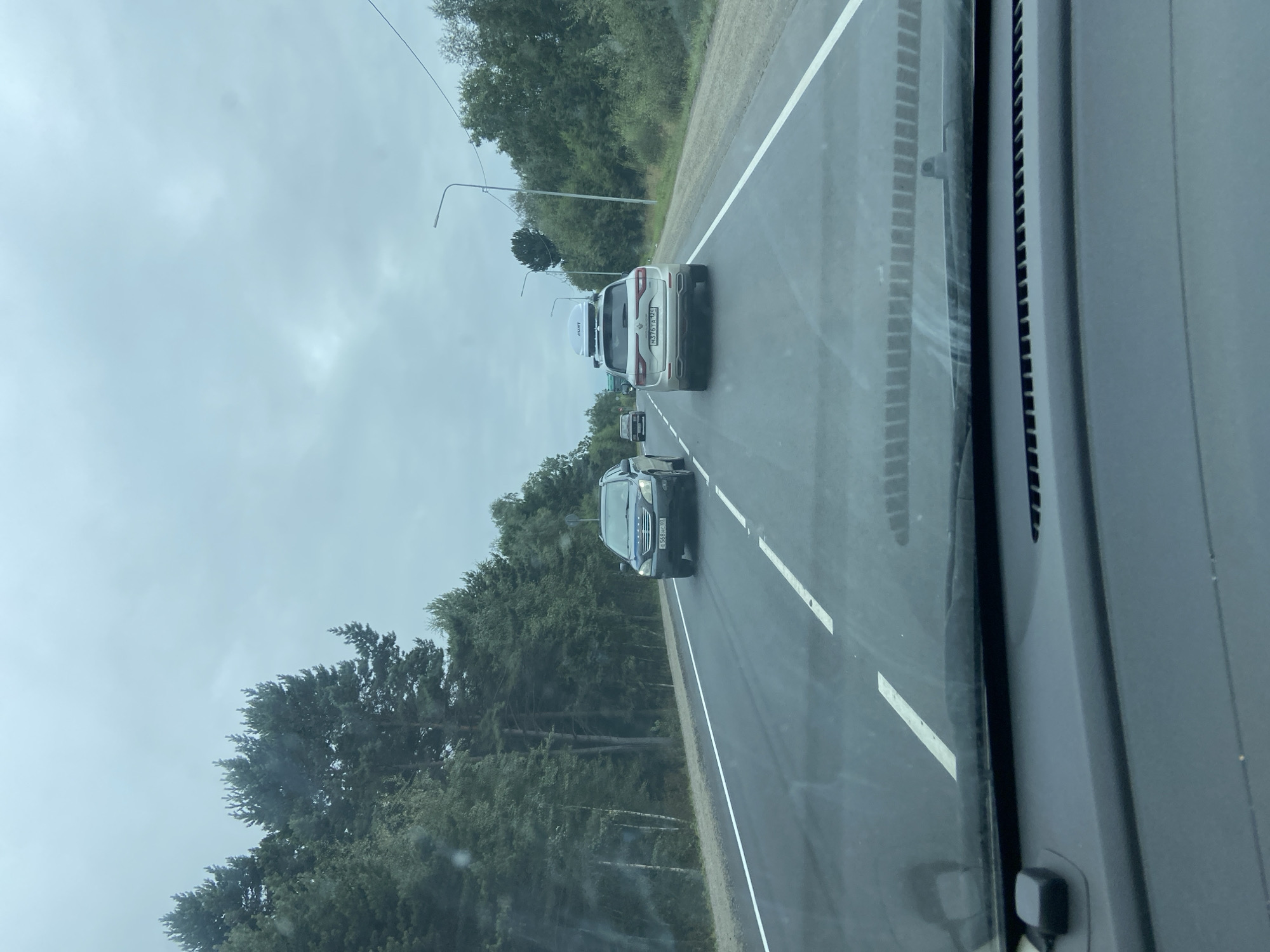 The image shows a view from inside a car driving on a two-lane road. The road is paved and has white lane markings. Trees line both sides of the road, and the sky is overcast.
In front of the car, there are several other vehicles on the road. The closest vehicle in front is a white SUV with a silver roof box. Behind it, a darker colored SUV is visible, and further ahead, a truck is partially visible. On the left side of the road, another dark-colored SUV is driving in the opposite lane.
The dashboard of the car from which the photo is taken is partially visible in the foreground, including a small black object mounted on the dashboard. The windshield is also visible, showing some reflections.
The scene appears to be a rural or suburban setting, given the presence of trees and the relatively empty road. The weather is likely cool or damp due to the overcast sky. The location is identified as Klyuyevka, Russia.