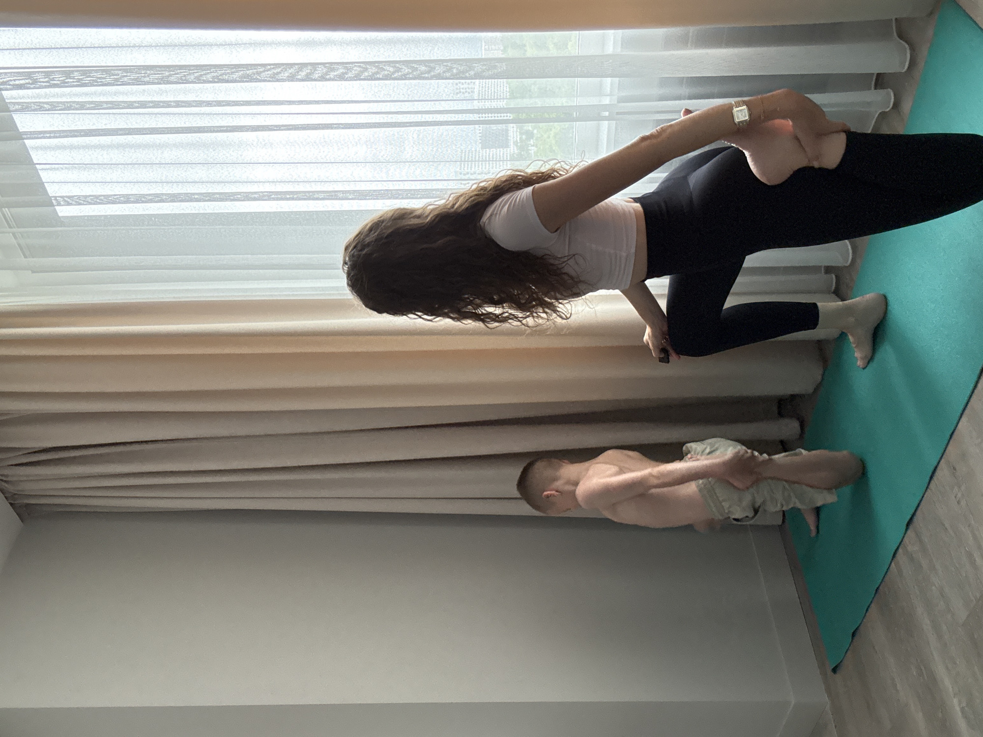 A mother and son are practicing yoga on a teal mat in a room with light gray walls and floor-to-ceiling curtains. The mother, with long curly brown hair, wears a white cropped t-shirt and black leggings, and is in a standing yoga pose, holding her foot behind her. Her son, with short brown hair, stands facing away from the camera in a similar pose, his left leg bent behind him. The room is lit by natural light streaming through sheer white curtains covering a large window. Buildings and trees are visible through the window.