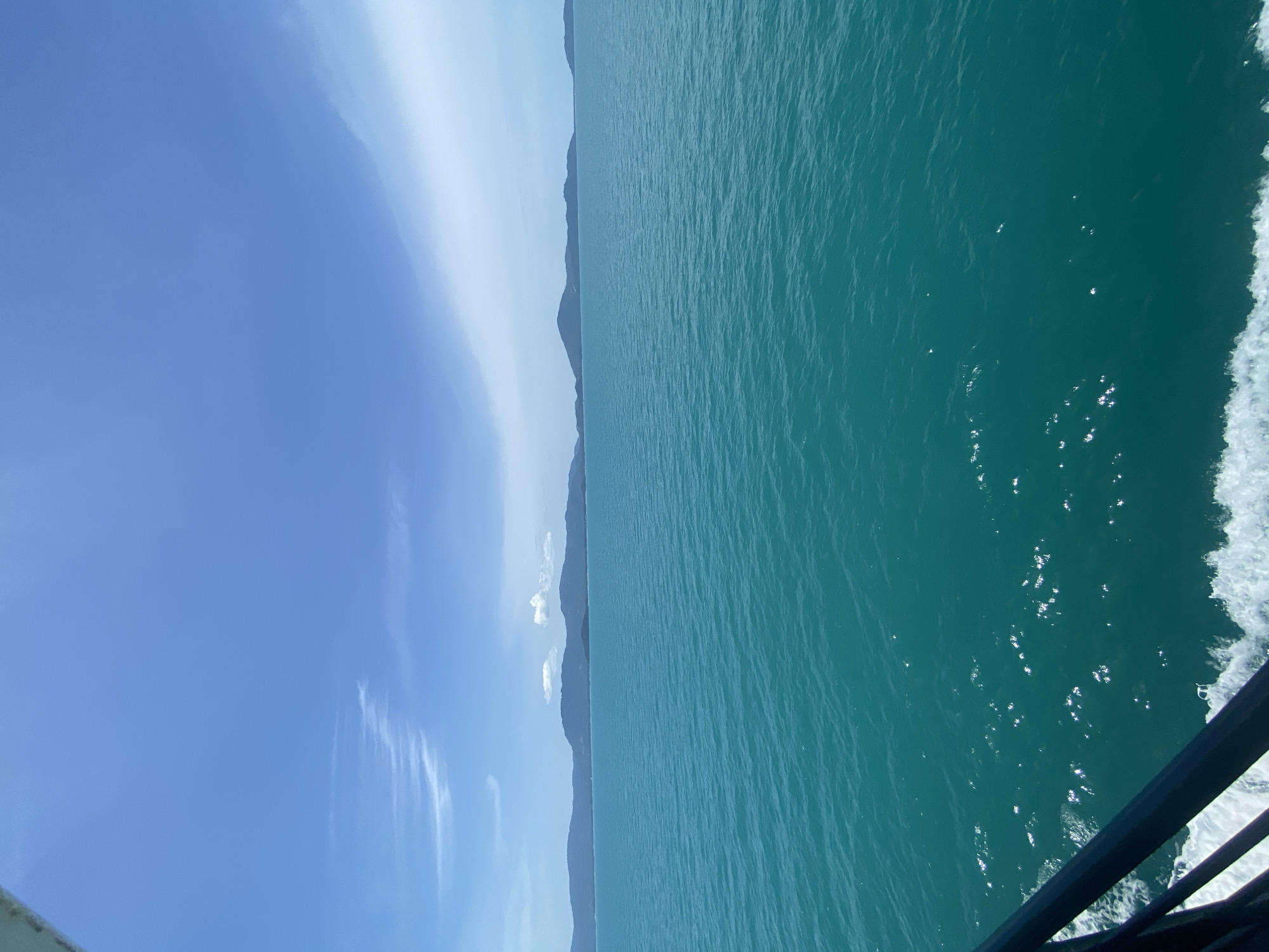 The image depicts a view from a ferry traveling on the ocean, likely near an island chain in Thailand. The water is a vibrant turquoise, with gentle waves creating sparkling reflections of the sun. In the distance, a range of lush green islands forms a natural horizon, some topped with scattered white clouds. The sky is a clear, bright blue, with wisps of thin clouds stretching across the expanse. In the foreground, a portion of the ferry's railing is visible, painted in a dark color, and a white wake churns behind the vessel, indicating movement. The scene suggests a bright, sunny day, typical of a tropical location.