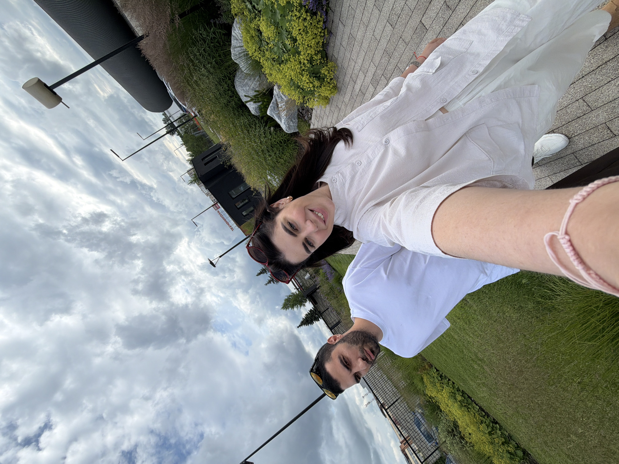 The image shows a selfie of a man and a woman taken outdoors. The woman, on the left, is wearing a white shirt and has long dark hair. She is smiling at the camera. The man, on the right, is wearing a white t-shirt and sunglasses. He has a beard and is looking towards the camera with his tongue sticking out. They are both smiling and appear to be in a playful mood.
They are standing in a park-like setting with lush green grass and bushes. In the background, there is a cloudy sky and some modern architecture, including a dark building with windows and a large cylindrical structure. There are also streetlights visible. The lighting suggests it is daytime, possibly late afternoon due to the quality of the light and the presence of clouds. The overall atmosphere is casual and joyful, indicative of a relaxed outing. There is a pink bracelet on the woman's wrist. No visible text is discernible in the image.