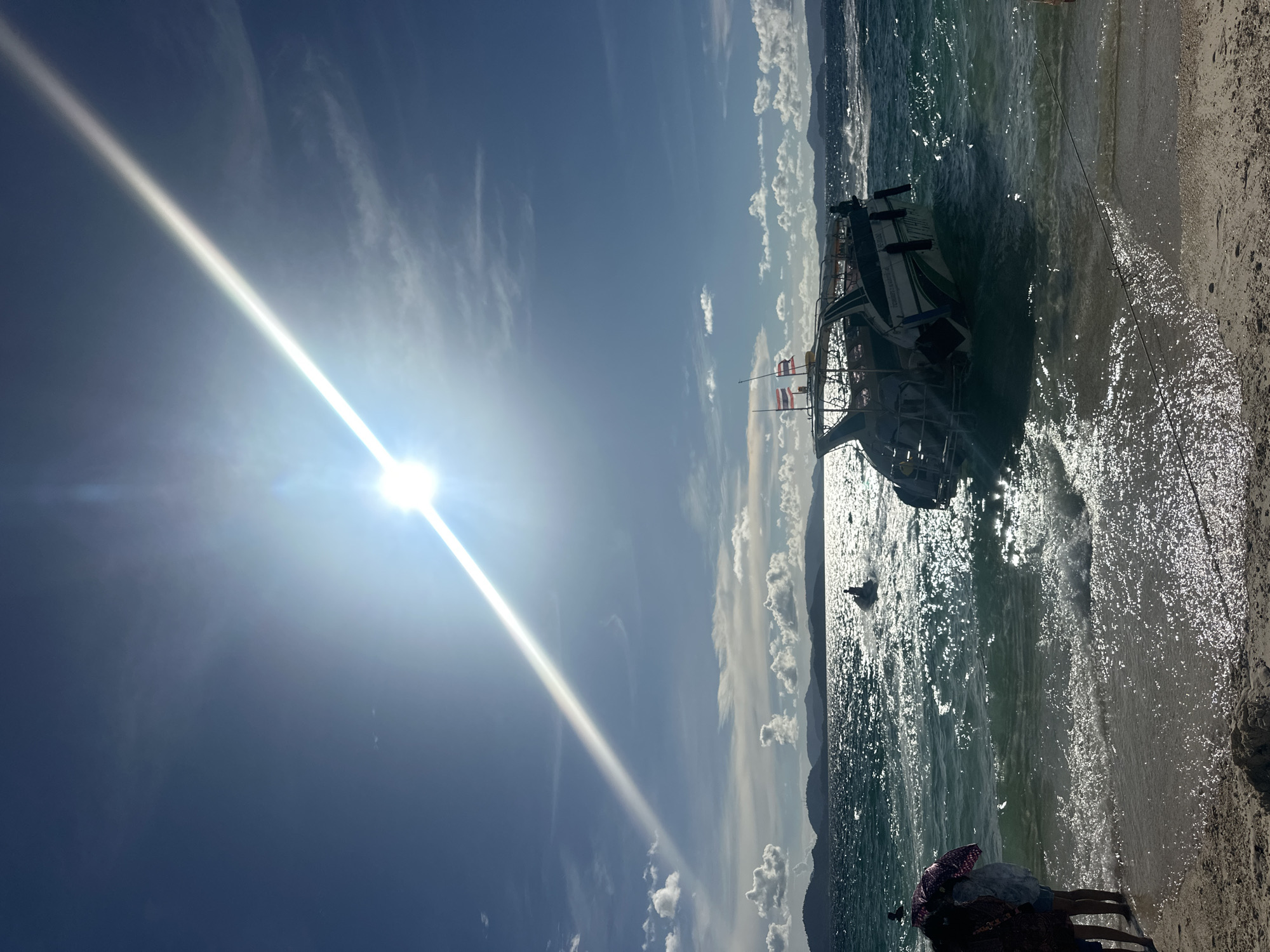The image depicts a vibrant daytime scene at a beach in Phru Nai, Thailand. The bright sun shines intensely, creating a strong beam of light across a clear blue sky dotted with fluffy white clouds. In the foreground, the edge of a sandy beach is visible, with gentle waves lapping at the shore.
The main object in the scene is a white and green boat, partially submerged in the shallow water near the beach. The boat appears to be a passenger vessel, possibly a tour boat, with multiple rows of seats and a canopy. Two small flags, red and white, are visible on the boat.
Several people are present. In the lower right corner, a small group of individuals are gathered on the beach, some partially obscured. One person is wearing a colorful patterned scarf. Further out in the water, near the boat, a person appears to be swimming or wading.
The overall mood is one of a sunny, leisurely day at the beach. The sparkling water and bright sunlight suggest warm weather and a pleasant atmosphere. The distant outline of mountains can be seen across the water, providing a scenic backdrop to the tropical setting. No specific text is legible in the image.