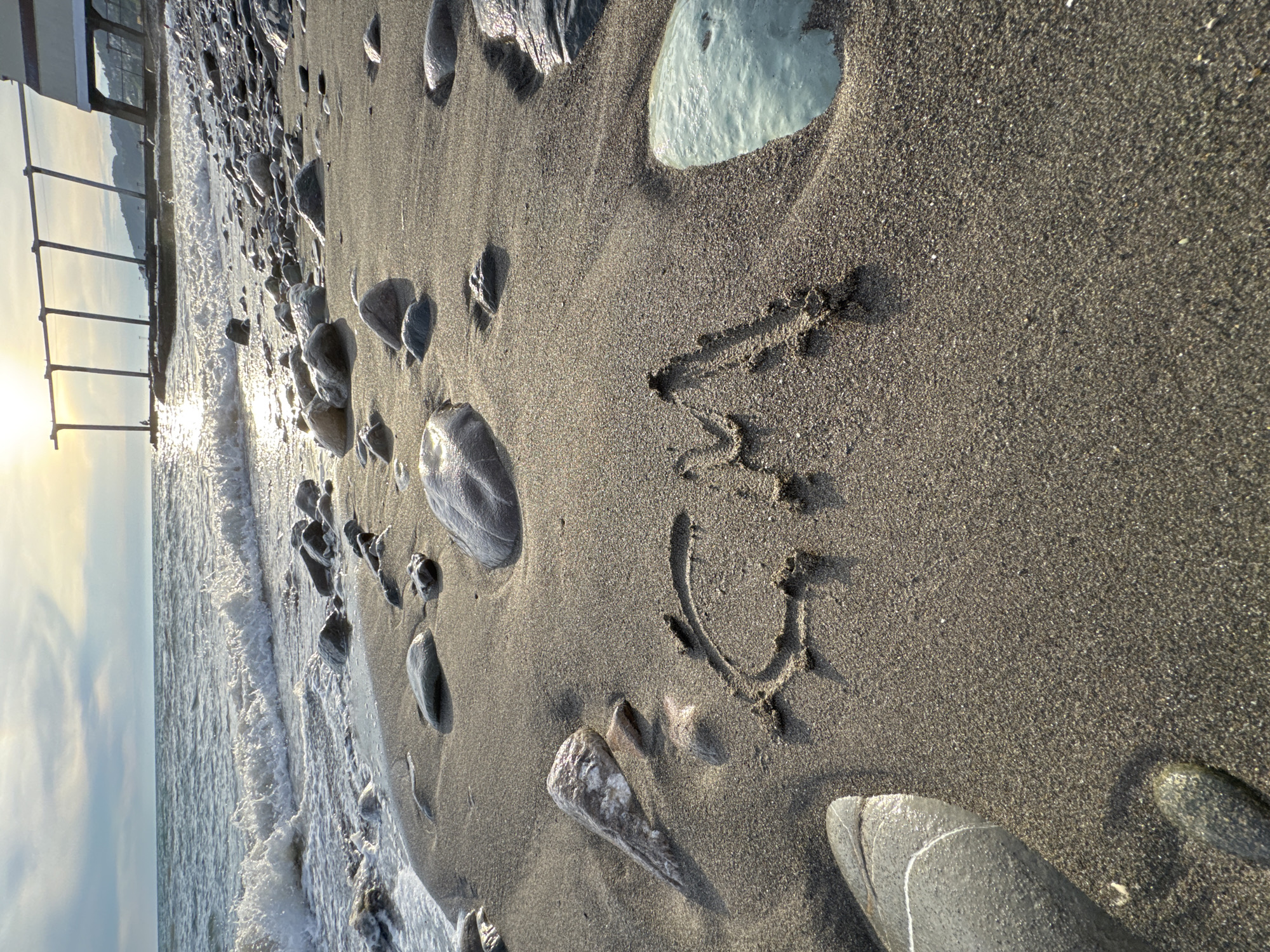 The image depicts a sandy beach in Sochi, Russia, with the sea in the background. The sky is partly cloudy with the sun setting, casting a warm glow on the scene. Waves are gently lapping onto the shore, and the beach is dotted with various smooth stones. In the foreground, the letters "CM" are written in the wet sand. A modern structure with elevated beams and a building is visible in the distance, along the coastline, suggesting an urban or resort area. The overall atmosphere is serene and picturesque, indicative of a late afternoon or early evening on the Black Sea coast.