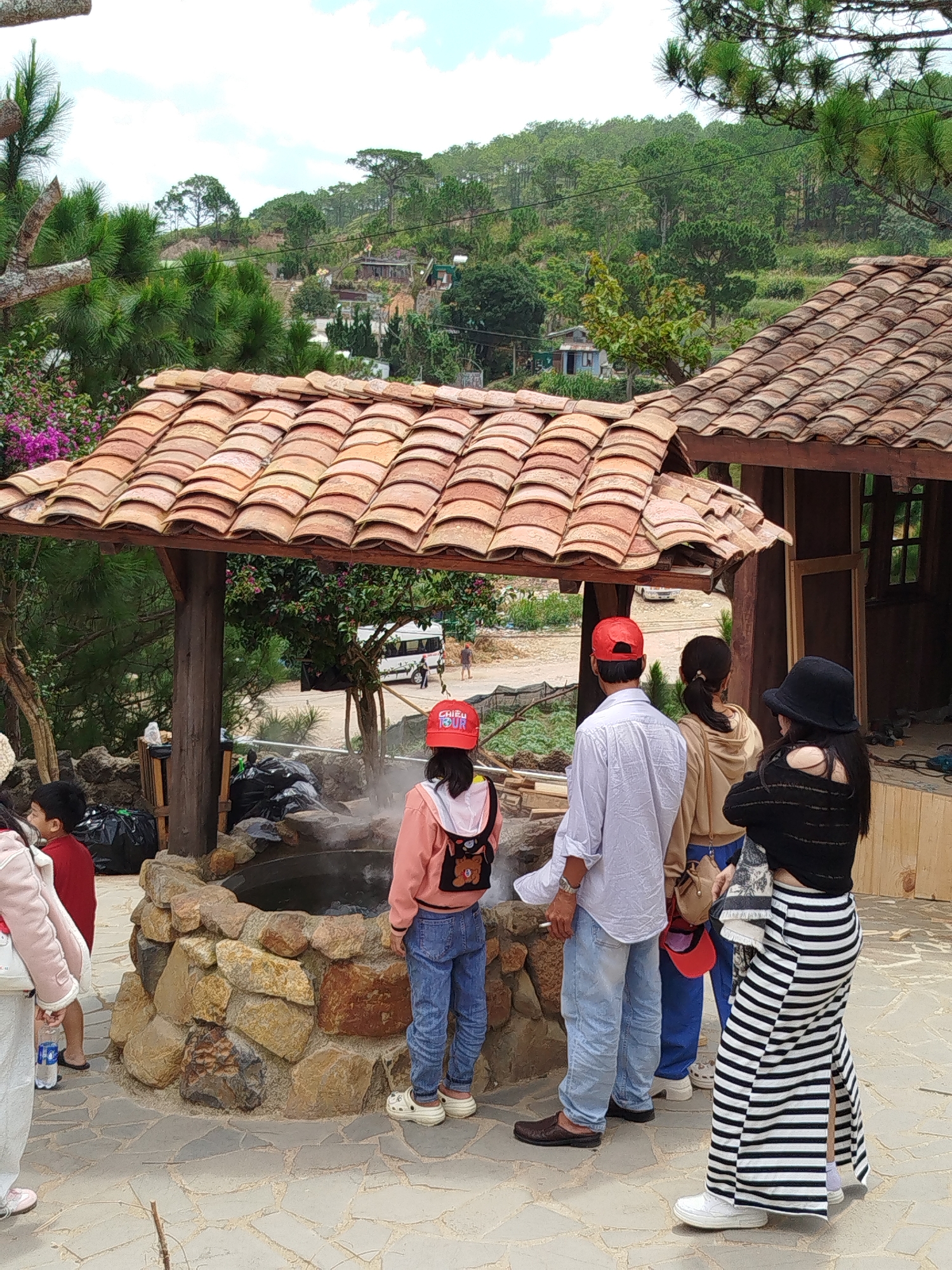 In the city of Xuân Thành, Vietnam, a group of individuals, including children, gathers around a steaming hot spring. The hot spring is contained within a circular structure built of rough-hewn stones. Overhead, a tiled roof supported by wooden posts offers shade. The surrounding environment features lush green hillsides dotted with trees and houses, suggesting a rural or mountainous area. A white van is visible in the background, parked on a paved road, with a few people standing near it. The people in the foreground are dressed casually, with some wearing hats and backpacks. The overall scene depicts a tourist attraction focused on a natural hot spring.