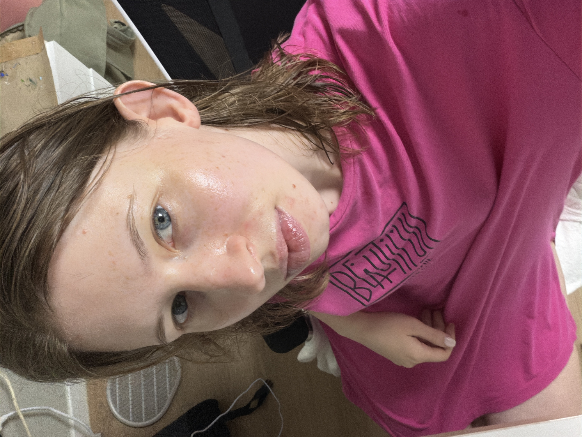 The media file is a close-up, rotated portrait of a young person with light skin, freckles, and blue eyes, wearing a bright pink t-shirt. Their hair is a medium brown and appears slightly damp. The person is looking upwards towards the camera, with a neutral expression. Some of the background is visible, including what appears to be a desk or workspace with art supplies. A small black fan is also visible on the floor. The lighting suggests it could be daytime. The setting is indoors, likely a home or studio, in Dongducheon-si, South Korea. A black graphic with wavy lines is printed on the person's t-shirt.