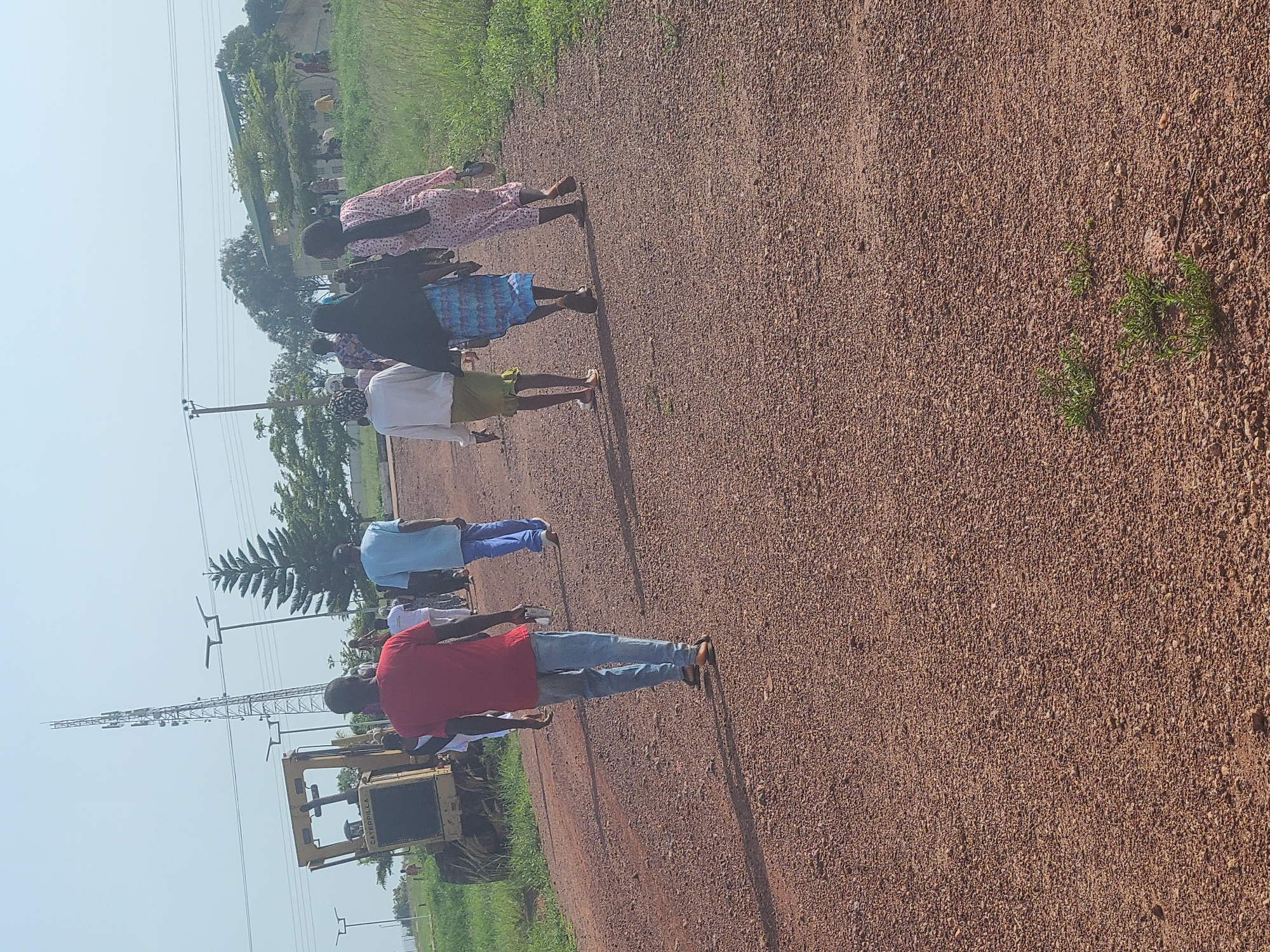 A group of people are walking away from the camera on a gravel road. The road is brown and dusty, with some small green plants growing on the sides. In the background, there is a large yellow caterpillar grader, a cell tower, and some trees. A few buildings are visible in the distance. The sky is a clear, pale blue. The location is Durumi, Nigeria.