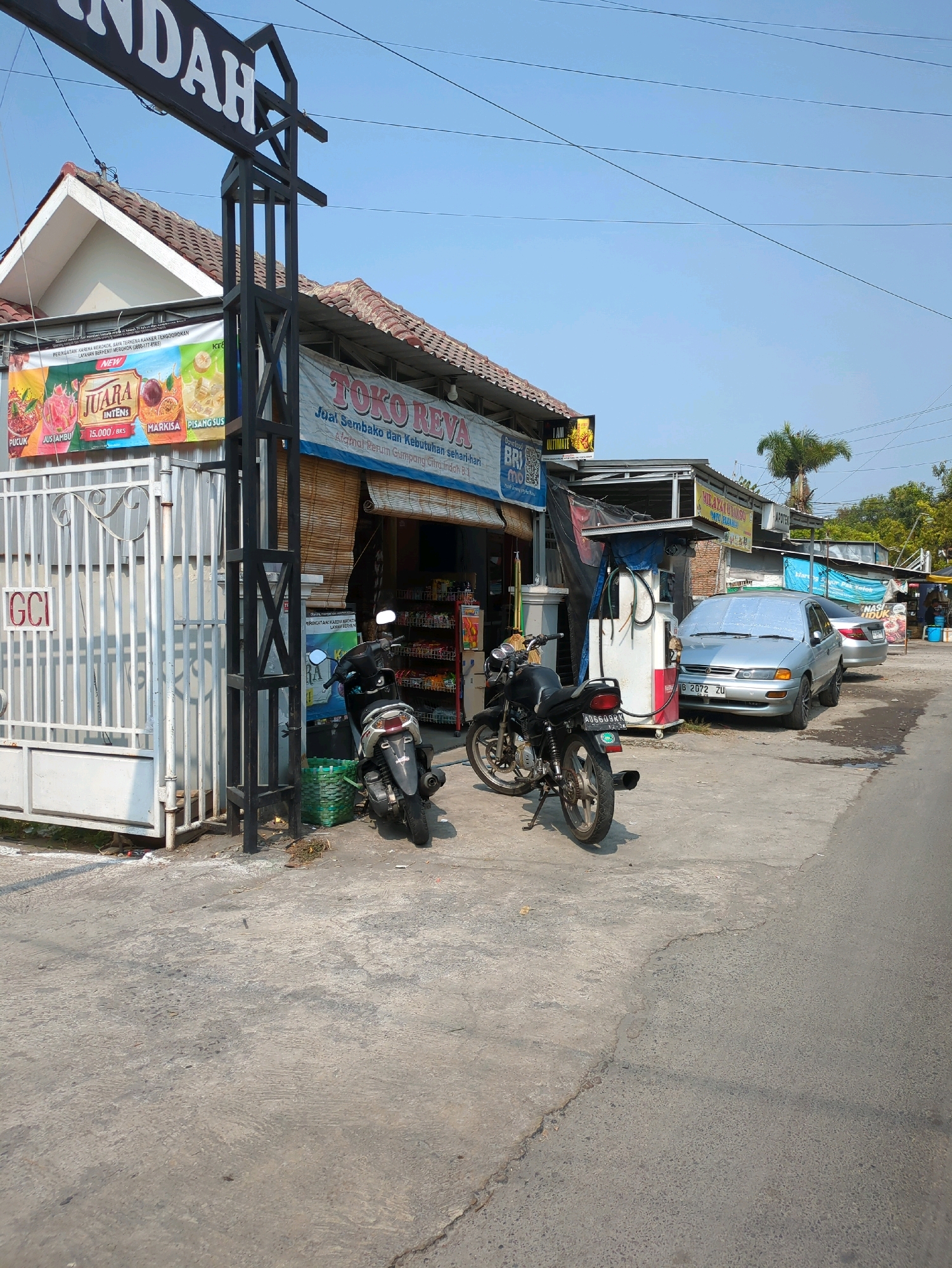 The image shows a street scene in Gumpang, Indonesia, on a clear, sunny day. In the foreground, a paved road transitions to a concrete area. A dark-colored motorcycle is parked on the concrete, with its kickstand down. To the left, a white metal gate with the letters "GCI" is visible.
Behind the gate, a building with a white exterior and a tiled roof is present. A banner above the building reads "INDAH." Adjacent to this building is a storefront labeled "TOKO REVA" with a bamboo-covered awning. Various items are displayed on shelves inside the store.
In front of the "TOKO REVA" building, a red and white gasoline pump stands. A silver sedan is parked next to the pump. Further down the street, another car is partially visible, and more storefronts line the road. Trees and power lines are present in the background. The overall impression is that of a small roadside business or a convenience store in a semi-rural or suburban area.