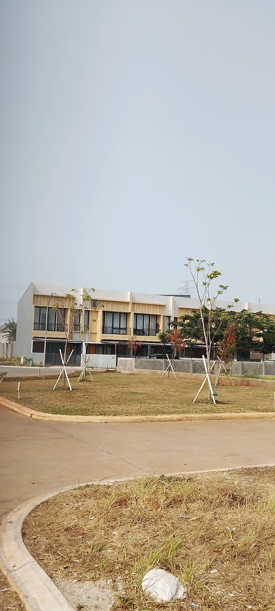 A perspective shot captures a pale sky over a modern, multi-unit building in Satriamekar, Indonesia. The building features a beige facade with dark-trimmed windows and balconies on its upper floors. In front of the building, young trees are planted in a grassy area, supported by wooden stakes. A paved road curves into the foreground, bordered by dry, patchy grass and a concrete curb. The scene appears to be a residential development or commercial complex under construction or newly built, with an air of stillness under the overcast sky.