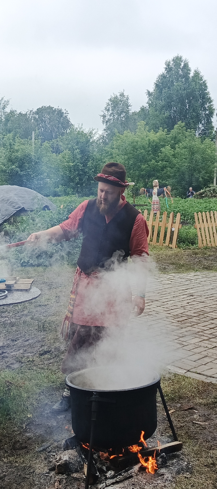 In Старые Быги, Russia, a man with a beard, wearing a brown hat with a red band and a dark vest over a red shirt, stands outdoors. He is stirring a large black pot filled with boiling liquid, which is generating a significant amount of steam. The pot is placed over an open fire, with visible flames licking at the bottom of the pot. The scene appears to be set in a rural or garden environment, with lush green trees and foliage in the background. A wooden fence and a scarecrow are visible further in the background, suggesting a village or agricultural setting. The ground is a mix of grass, dirt, and a paved area with interlocking bricks. The sky is overcast, indicating a cloudy or possibly rainy day. The overall mood seems to be one of outdoor cooking or a traditional gathering. There is no visible text in the image.