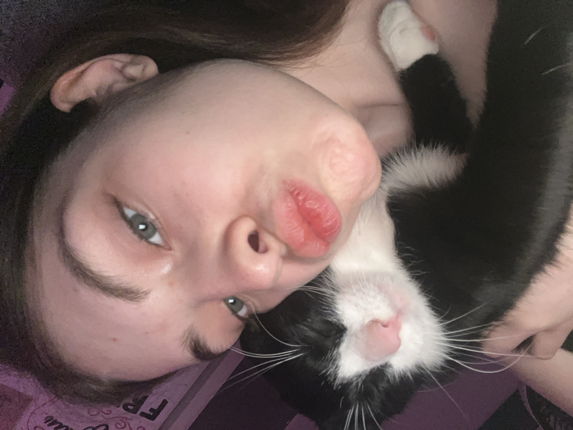 A young person with fair skin and brown hair is lying down, posing for a selfie with a black-and-white cat. The person is making a kissing face, and the cat is snuggled close to their face. The cat has a pink nose and white whiskers. The background is blurred. This scene is not set in Saint Petersburg, Russia.
