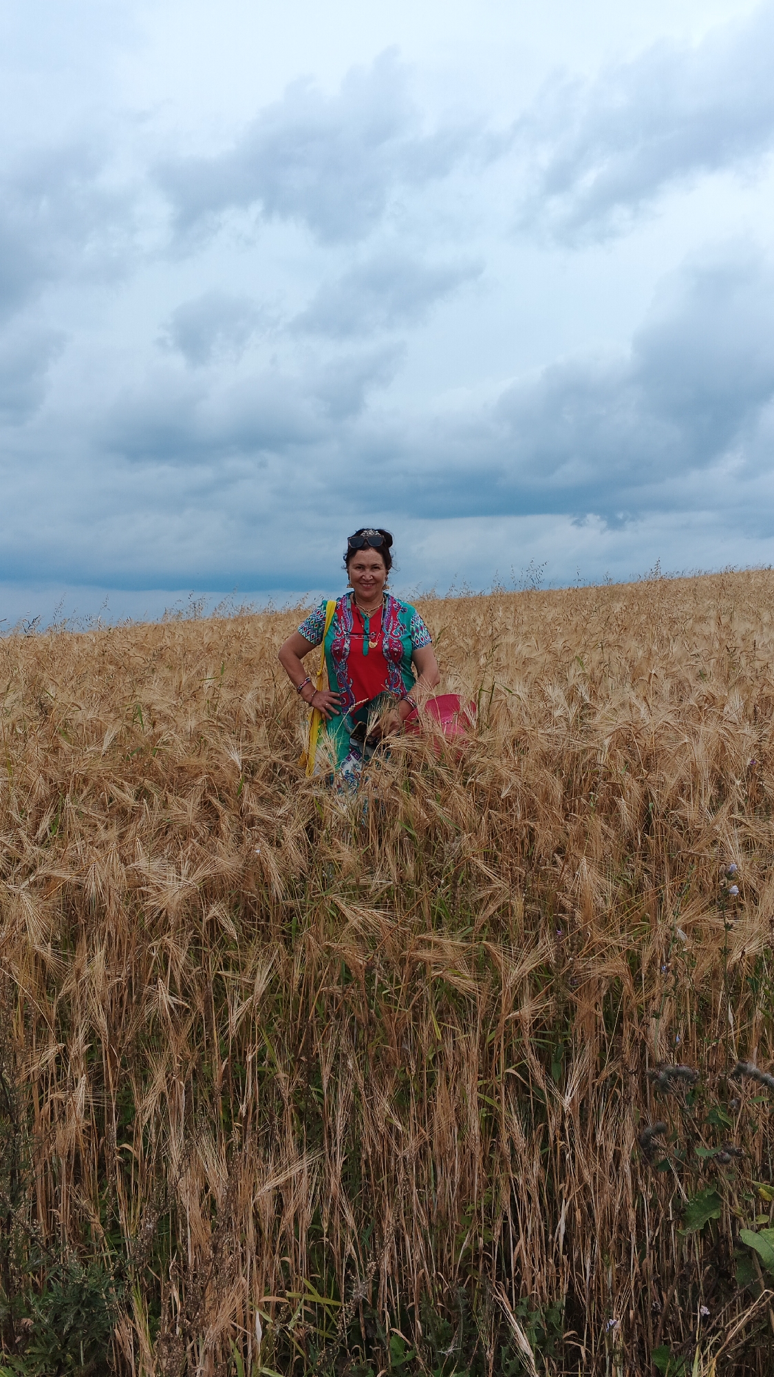 A woman stands in a wheat field under a cloudy sky. The woman is wearing a brightly colored outfit with a red top and a patterned green and white overlay. She has dark hair pulled up, sunglasses on her head, and is wearing jewelry. She is positioned in the center of the frame, with her hands on her hips and a slight smile. To her right, a pink bag is visible. The wheat stalks are golden brown and reach up to her waist. The background shows a slightly hilly landscape covered in more wheat, leading to a cloudy sky with patches of lighter blue. The location is identified as Podgornoye, Russia.