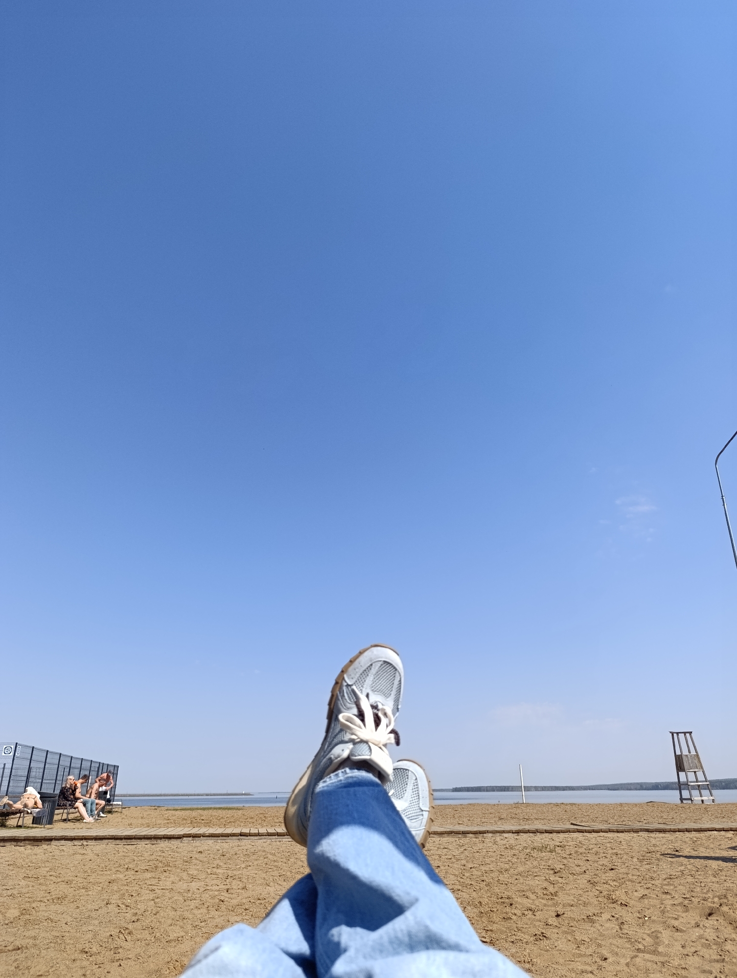 Here is a description of the image:
The image shows a first-person view of someone relaxing on a sandy beach under a clear blue sky. The person's legs, clad in blue jeans and light-colored sneakers, are crossed in the foreground. In the background, there is a body of water, likely a lake or river, and a shoreline with some sparse trees in the distance. The beach is sandy, and there are some objects on it, including a lifeguard stand and a fence with people sitting or lying down. The location is Chaikovsky, Russia.