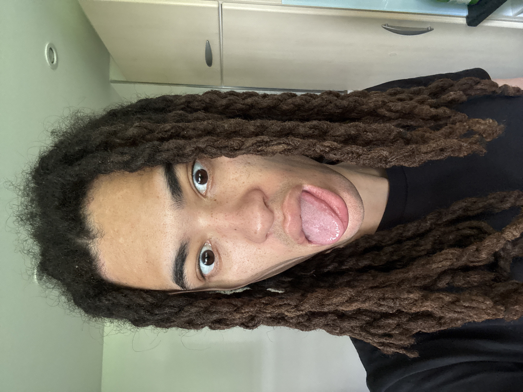 A man with brown dreadlocks, wearing a black shirt, sticks his tongue out at the camera. He is indoors, in what appears to be a home or apartment with a wall and cabinets visible in the background. There is no verifiable evidence to conclude that this image was taken in Les Ulis, France.