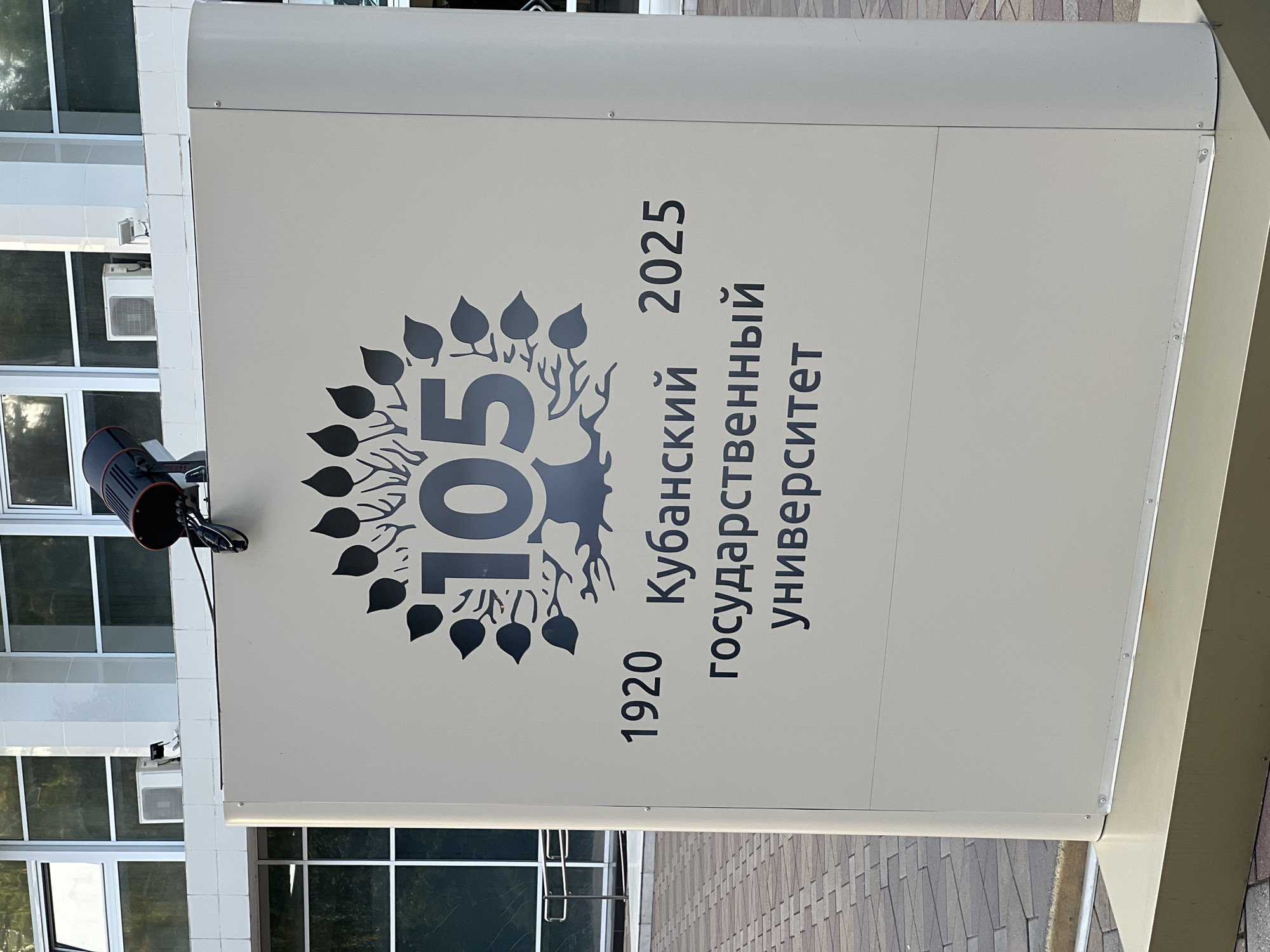 This is a photograph of a large, freestanding sign outside a building. The sign is light beige and has dark gray text and graphics. At the top, there is a stylized tree with leaves and the number "105" superimposed on it. Below the tree, the text reads "1920 Кубанский 2025 государственный университет". The sign is located in front of a modern building with large windows and white architectural elements. The ground in front of the sign is paved with light-colored brickwork. The sign is likely located at the Kuban State University in Krasnodar, Russia, commemorating its 105th anniversary from 1920 to 2025. A black industrial lamp is mounted on top of the sign.