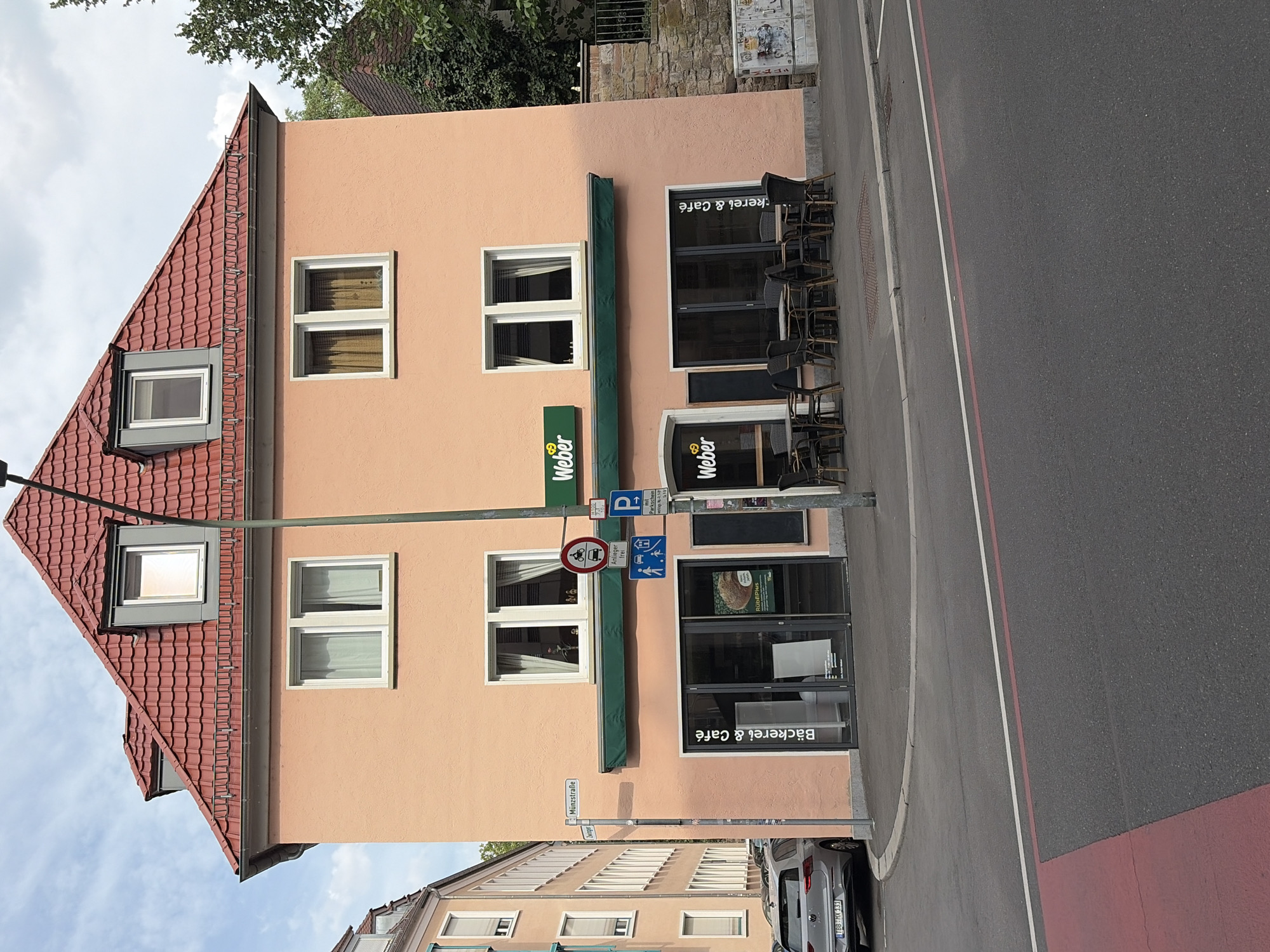 This image shows the exterior of a multi-story building in Würzburg, Germany, with the street and sidewalk in the foreground. The building is painted a peach color and has a red tiled roof with dormer windows. On the ground floor, there are storefronts for a bakery and cafe, identified by "Bäckerei & Café" and "Weber" signage. Outdoor seating with tables and chairs is set up in front of the cafe. Several street signs are visible, including "Münzstraße" and "Zwinger," indicating the location. A silver Volkswagen SUV is parked on the street. The sky is cloudy.