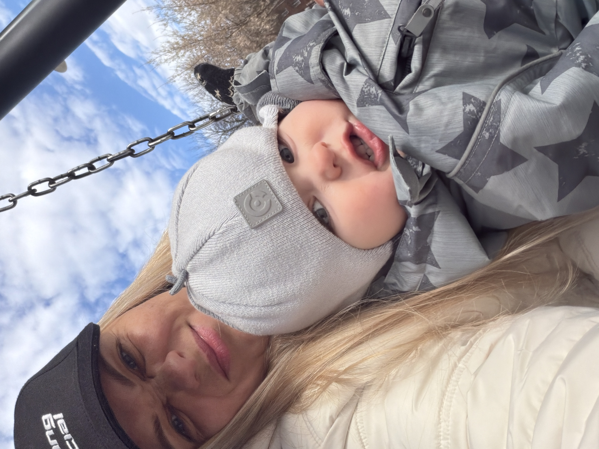 This close-up outdoor photograph captures a woman and a young child, likely a toddler, seemingly engaged in play on a swing. The image is rotated 90 degrees counter-clockwise, giving an unconventional perspective looking upwards towards the sky.
In the foreground, a woman with blonde hair is visible, wearing a black baseball cap with white text that appears to read "LCI Build" (the rest of the word is obscured). She has light makeup and a subtle smile. Her attire includes a light-colored, possibly off-white, quilted jacket.
Above her, the child is suspended upside down, looking directly at the camera with wide blue eyes and an open mouth, conveying a sense of surprise or playfulness. The child is dressed warmly in a light grey knit hat featuring a square patch with a circular logo, and a grey winter jacket adorned with a pattern of darker grey stars. A dark, patterned glove or boot is partially visible near the top left.
A heavy metal chain, typical of a swing set, stretches diagonally across the frame, connecting to an unseen swing seat. A dark metal bar, part of the swing structure, is also visible at the top left.
The background consists of a bright blue sky with scattered white clouds and the bare branches of deciduous trees, suggesting a clear day in late autumn, winter, or early spring. The scene depicts a cheerful interaction, possibly a parent swinging their child at a playground.