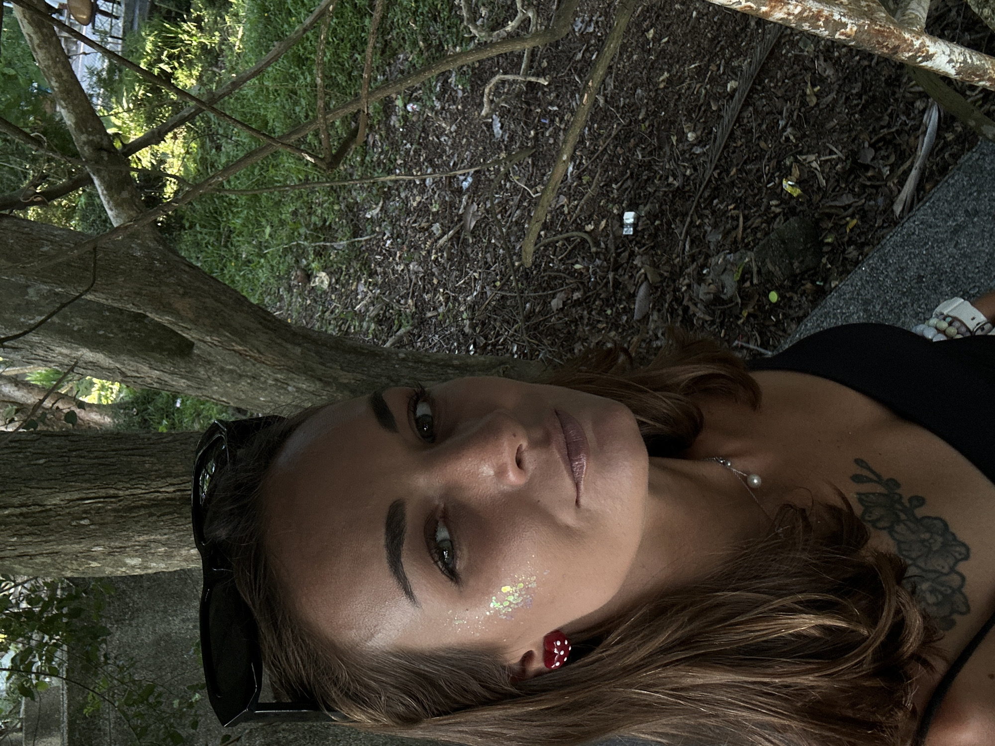 The image shows a close-up, low-angle shot of a young woman lying down outdoors. She is wearing a black top and a pearl necklace. Her hair is long and brown. She has glitter on her cheek and a red earring that looks like dice. There is a tattoo of flowers on her shoulder. In the background, there are trees, foliage, and debris on the ground. The lighting suggests it is daytime, possibly late afternoon, with sunlight filtering through the trees. The setting appears to be a natural, possibly wooded area. The woman's expression is relaxed and her gaze is directed upwards and to the side. The image is oriented vertically, as if the camera was held upright but the subject is lying down such that the top of her head is to the left. There is no visible text in the image.