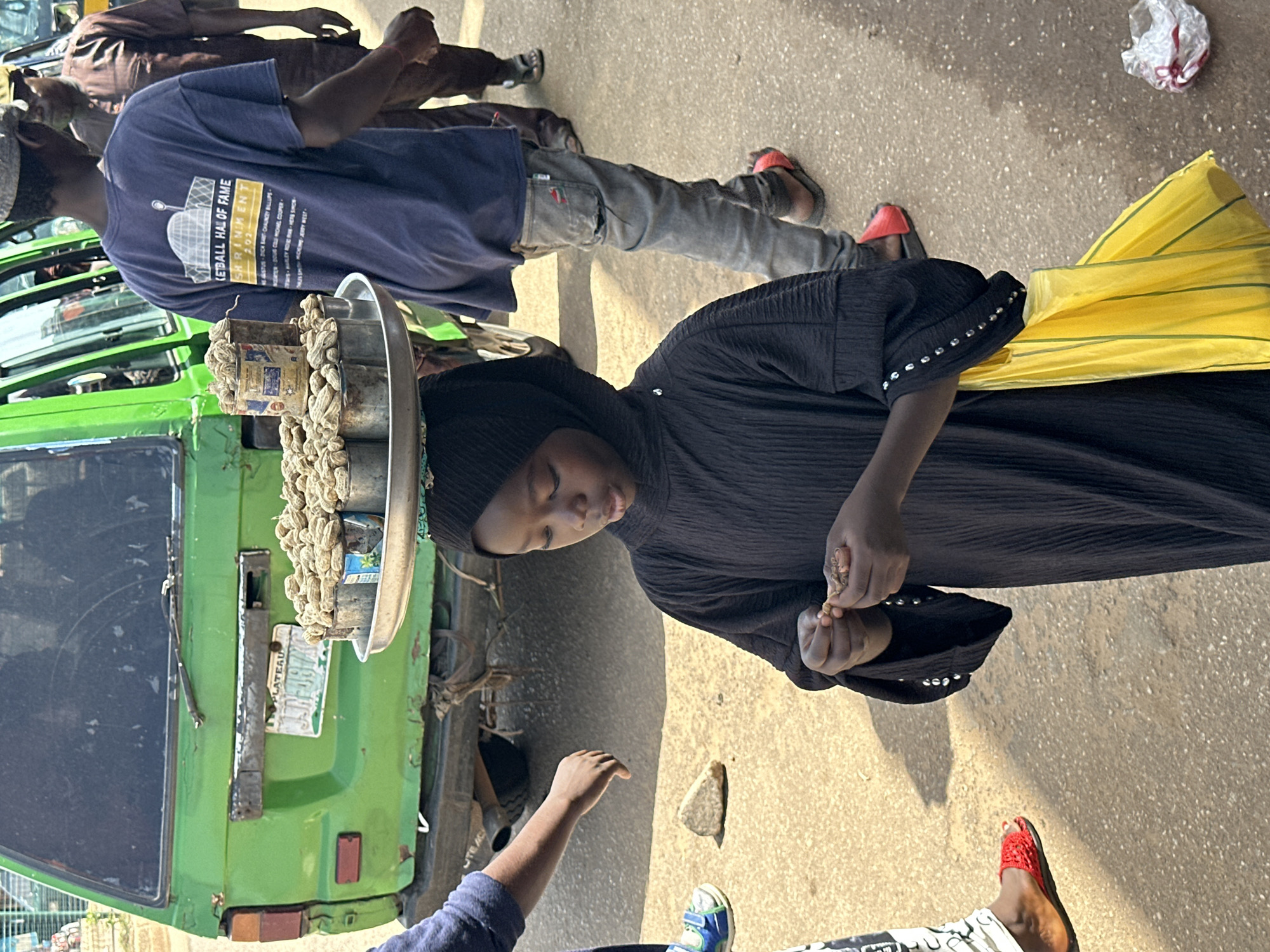 In Bukuru, Nigeria, a young girl wearing a black hijab and abaya balances a tray of peanuts on her head. She is dressed in a black abaya with beaded cuffs and holds a yellow bag in her right hand. The girl is in the process of shelling a peanut with her left hand. She stands on a paved street next to a bright green van. A man wearing a blue t-shirt with "Basketball Hall of Fame" text and grey jeans stands behind her, to her right. Further to the right, another man in a brown traditional outfit walks away from the camera. The background shows the rear of the green van, with a license plate that reads "PLATEAU / JHR-097X". The scene appears to be a street market or roadside setting with other pedestrians visible in the distance. The ground is a mixture of asphalt and dirt.