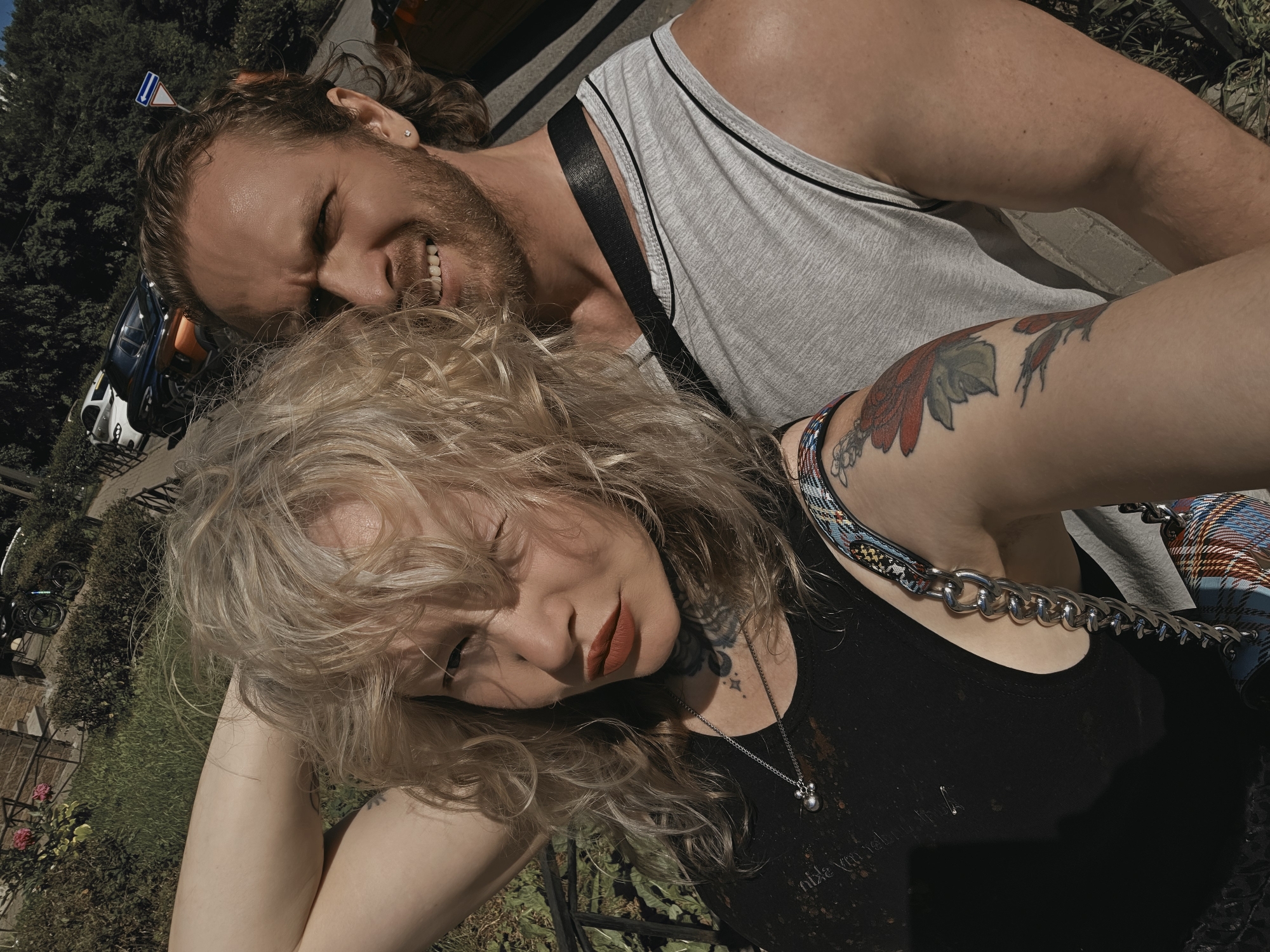 This is a selfie of a couple in a park in Saint Petersburg, Russia. The woman, on the left, has blonde, curly hair and is wearing a black tank top with a silver necklace and red lipstick. She has tattoos on her neck and arm. The man, on the right, has brown, curly hair and a beard. He is wearing a grey tank top. In the background, there are trees, bushes, and a street with cars. The sun is shining, casting shadows.