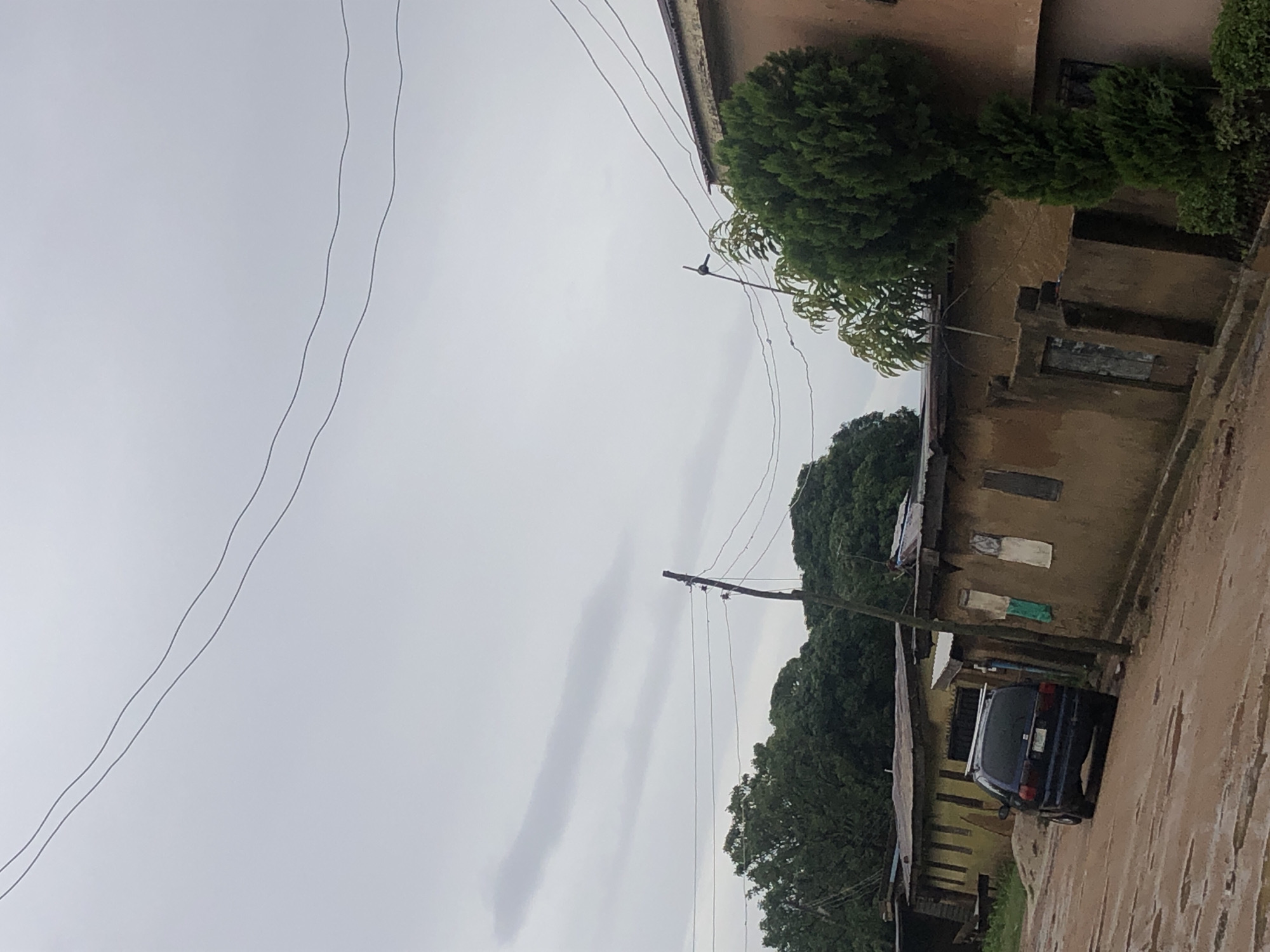 The image depicts a residential street scene in an urban environment. The road is unpaved and appears to be muddy, with puddles of water indicating recent rainfall. A dark-colored minivan is parked on the left side of the road, facing away from the camera. The street is lined with buildings constructed from earth or mud-brick, with corrugated metal roofs. These buildings are low-rise and appear somewhat weathered.
On the right side of the image, a tall, bushy evergreen tree is prominently featured. Behind the buildings, dense green foliage suggests the presence of trees and vegetation. Overhead, power lines crisscross the sky, supported by wooden poles. The sky is overcast with gray clouds, indicating a cloudy or potentially rainy day. The overall impression is of a humble, possibly developing, urban or semi-urban neighborhood.