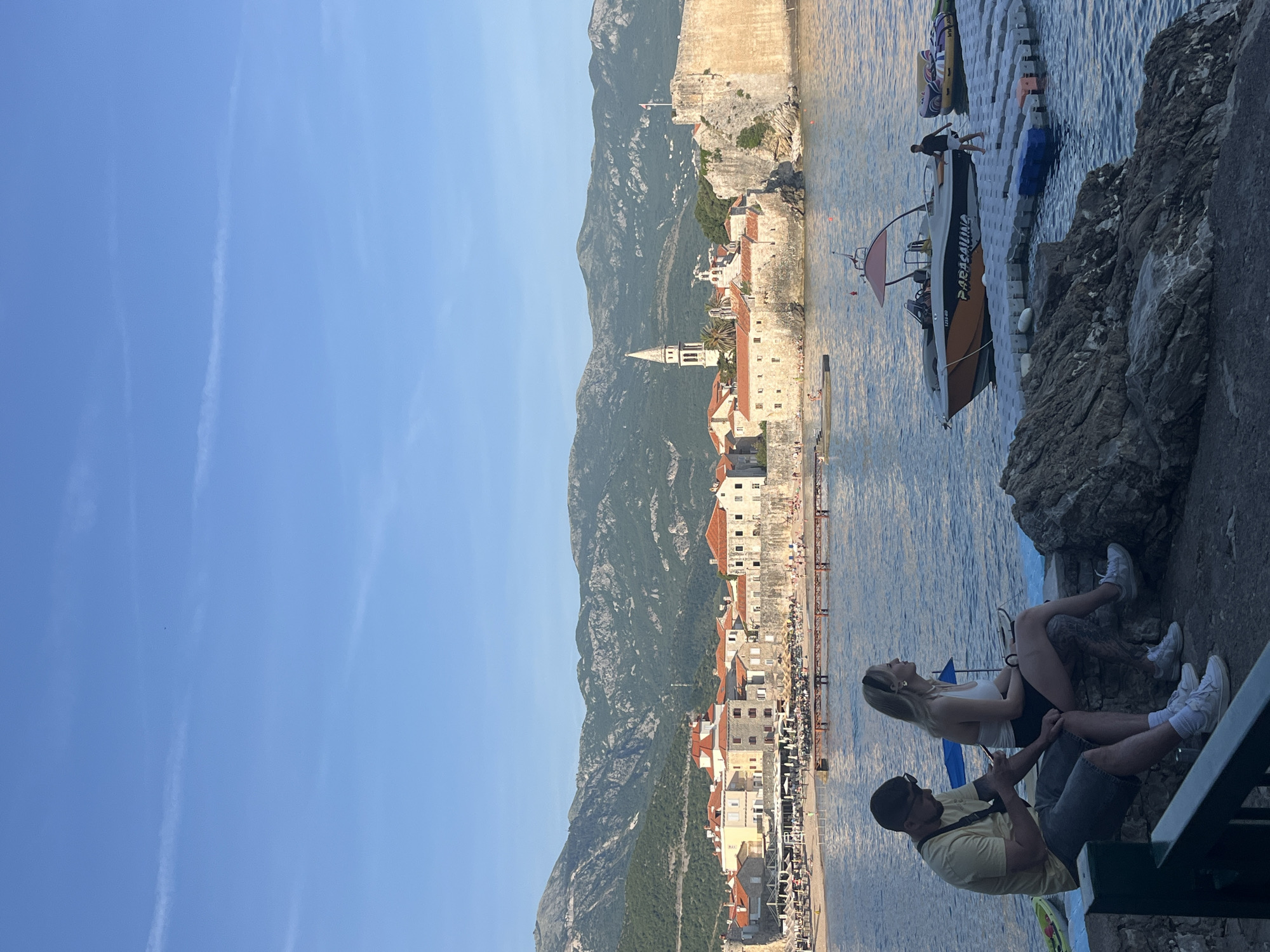 This media file captures a panoramic view of a vibrant coastal town, identified as Budva, Montenegro, nestled against towering, rugged mountains under a clear blue sky. The scene is illuminated by bright sunlight, suggesting daytime.
In the immediate foreground, two individuals are seated on a rocky outcrop by the water's edge. A woman with blonde hair, wearing a white top and dark shorts, is looking towards the left side of the frame. Beside her, a man with a beard and sunglasses, dressed in a yellow t-shirt and grey shorts, is focused on a mobile phone in his hands. Steps or a path are visible leading down near their seating area.
Further out on the calm, blue water, a small motorboat is docked next to a floating platform. A person in a dark top is standing on the front of the boat. The boat has the word "PARASAILING" prominently displayed on its side, along with a partial registration number like "OB-1215RD." An inflatable water tube or raft is also visible near the platform, indicating water recreational activities.
The mid-ground features the charming architecture of the town, characterized by light-colored stone buildings with distinct terracotta roofs. A prominent bell tower rises from one of the structures. A fortified stone wall runs along the coastline, separating the town from the sea, with a flag visible atop it. A long pier extends into the water from the town's edge.
The background is dominated by a majestic range of green and grey rocky mountains that provide a dramatic backdrop to the coastal settlement. The sky above is a brilliant azure, with faint, white contrails streaking across it, suggesting good weather conditions. The overall atmosphere is one of leisure and scenic beauty.