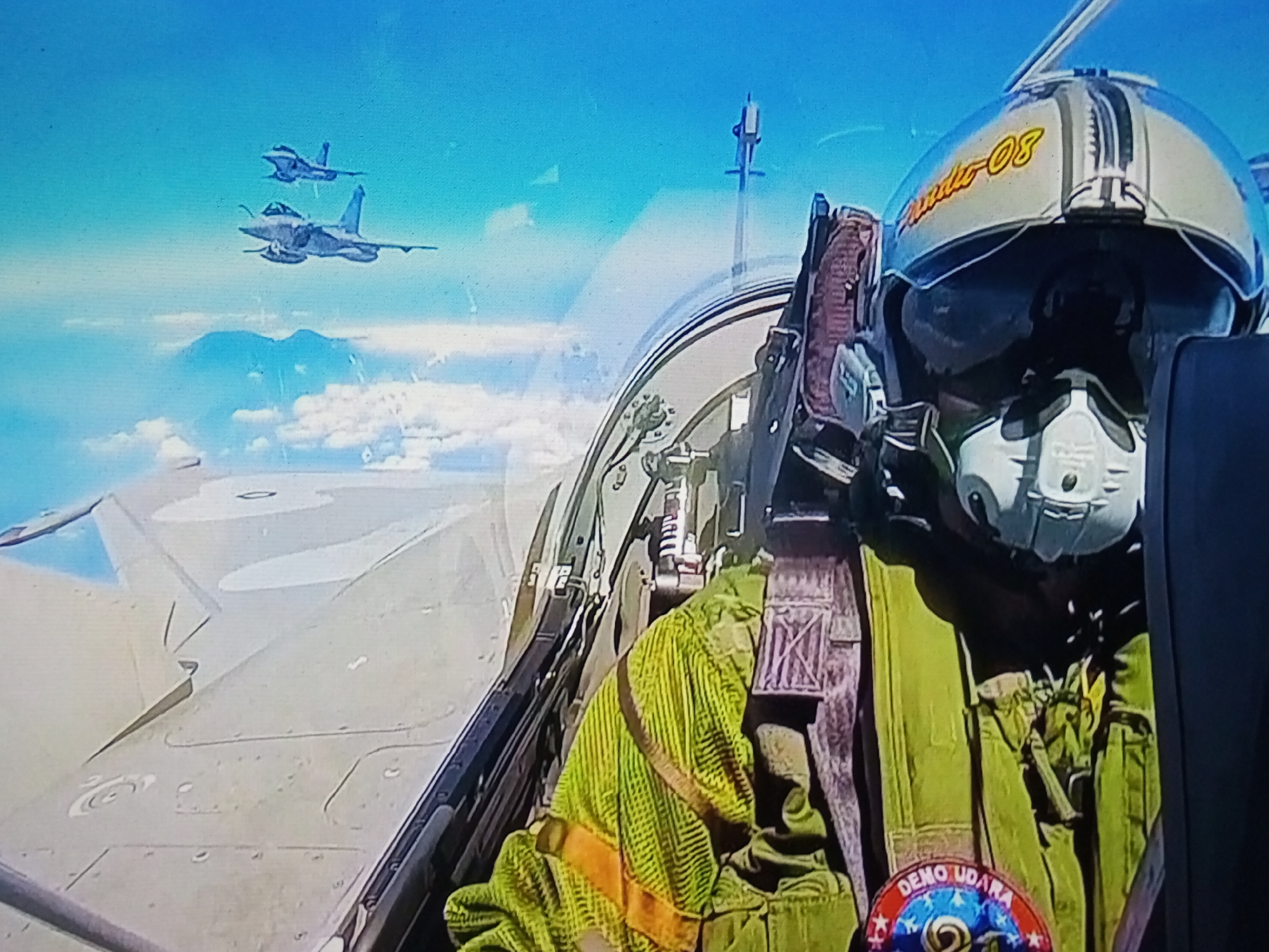 The image depicts an interior view from the cockpit of a fighter jet in flight, with a pilot wearing a helmet and flight suit visible in the foreground. Two other fighter jets are seen in formation against a clear blue sky and scattered clouds in the background. The pilot's helmet is white with yellow and black accents, and the inscription "NIDA-08" is visible on the side. The flight suit is green with orange straps. A patch is visible on the pilot's leg with the text "DEMO UDARA". The overall scene suggests an airshow or military exercise taking place in the skies above Banyudono, Indonesia.
