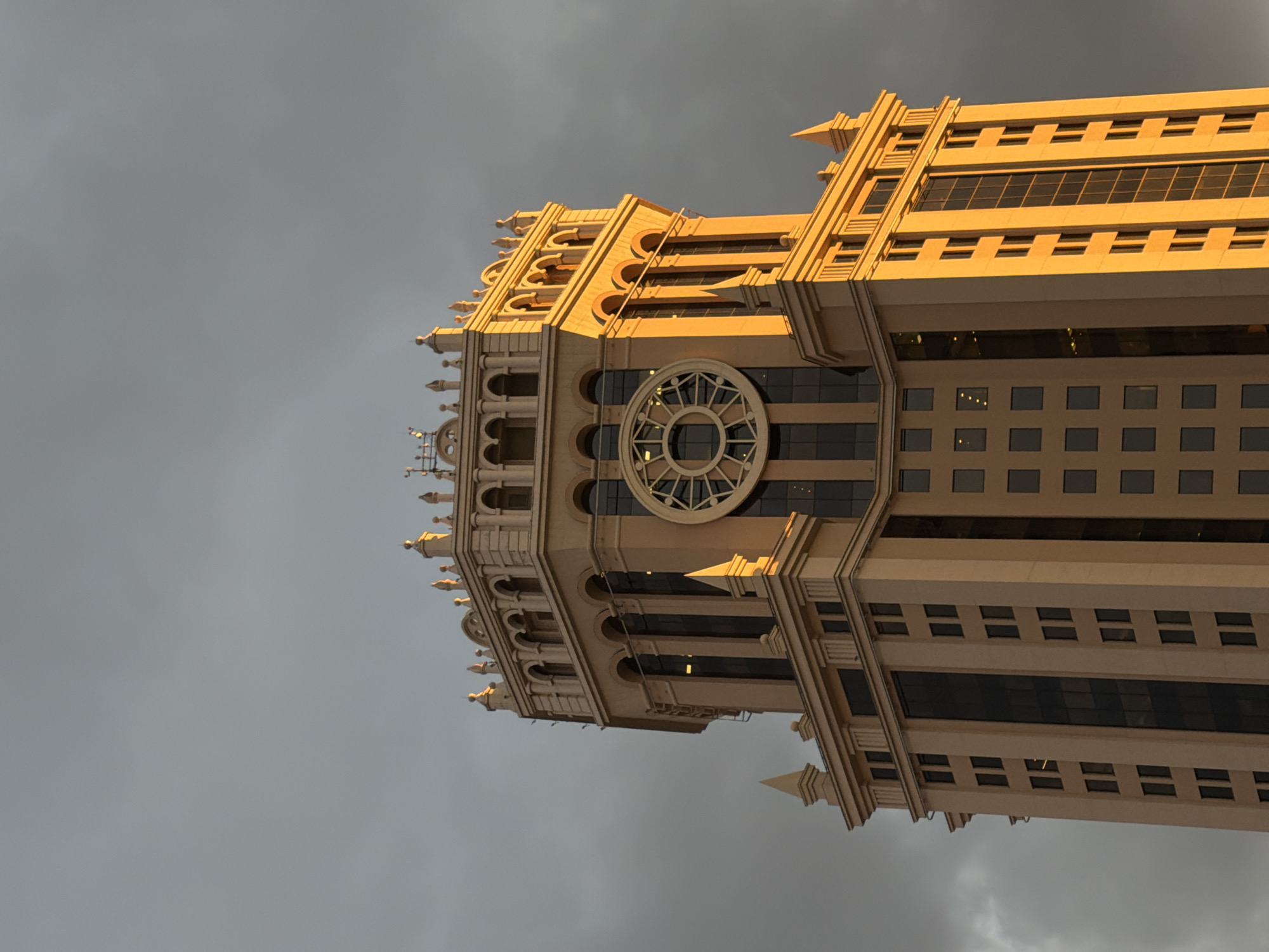A striking photograph captures an ornate, multi-storied building, oriented horizontally across the frame. The structure boasts a light-colored, possibly beige or light yellow, facade that is intensely illuminated by what appears to be the golden light of either sunrise or sunset. Its architecture is grand and intricate, featuring numerous decorative elements such as stepped setbacks, rounded arches, balustrades, and pointed spires or finials along its cornices. A prominent circular window, reminiscent of a rose window, with an elaborate spoked design, is visible near the center of the building's length. Below and above this circular window, and across other sections, are rows of rectangular windows, some appearing dark and reflective, others showing internal light sources. The building stands in dramatic contrast against a dark, overcast sky filled with heavy grey clouds, suggesting stormy or brooding weather. There are no people visible, nor any discernible text. The image's composition and lighting evoke a sense of majesty and drama.