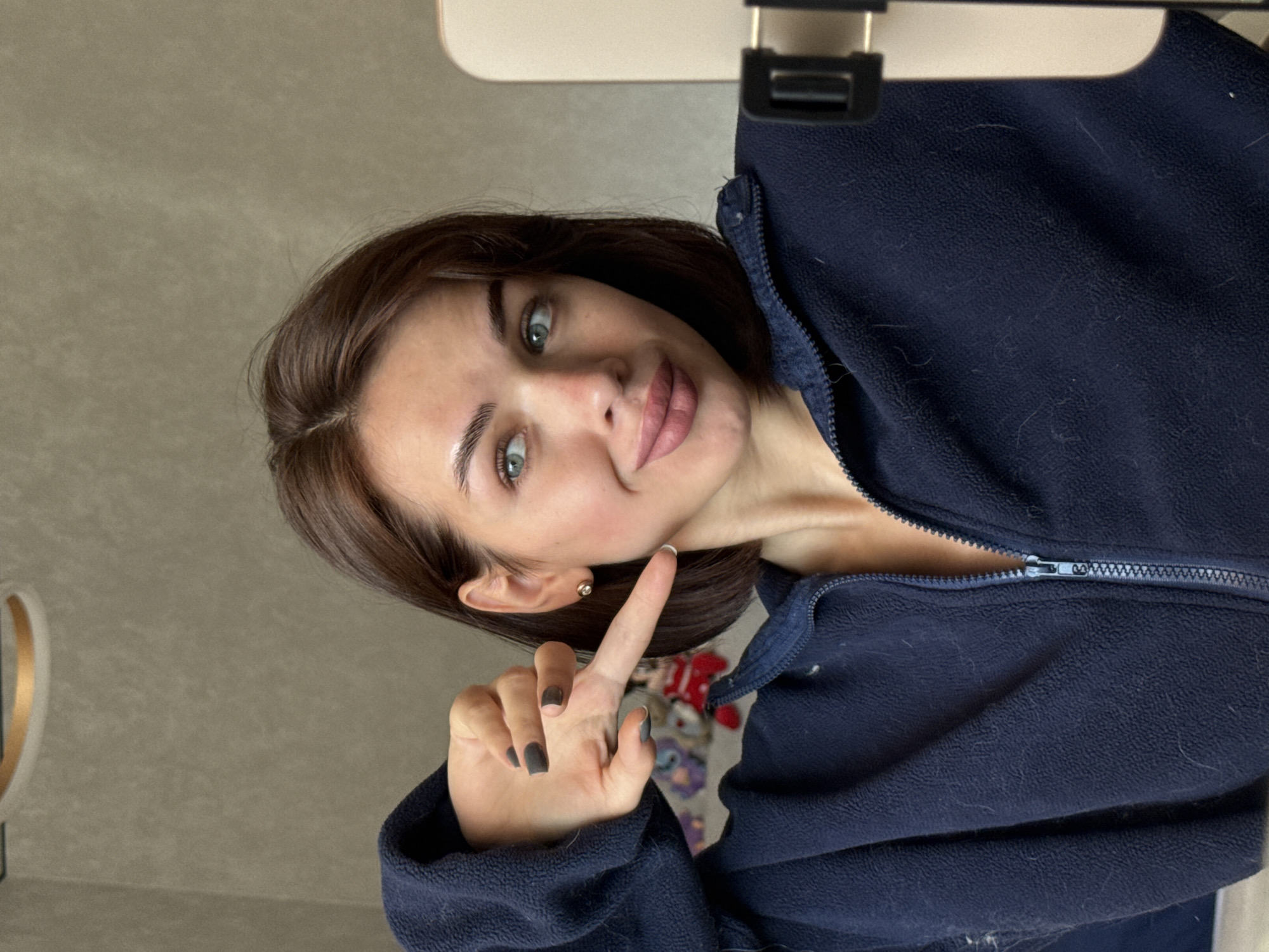 A young woman is holding up her index finger toward the camera. She has brown hair and green eyes. She is wearing a dark blue fleece jacket that is partially zipped. Her fingernails are painted a dark color. The background appears to be a room with a gray wall. The jacket is hanging on a rack. A blurred pattern can be seen between the jacket and the woman’s hand. It is not possible to ascertain the location as Лобаново, Russia from the image.