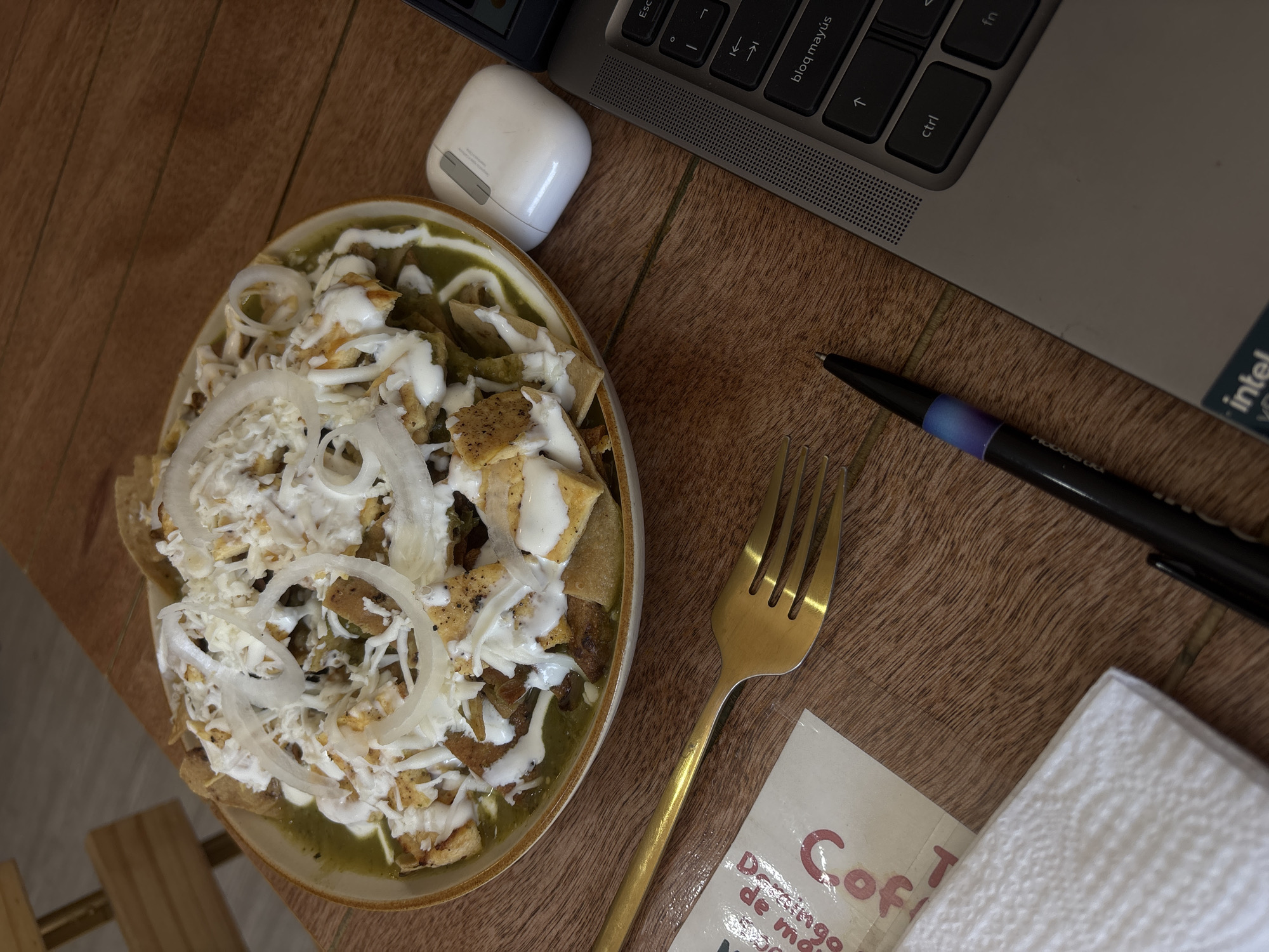 A high-angle shot shows a table setting with food and various items, potentially in Unknown, Unknown. A plate of Chilaquiles is heavily topped with cheese, cream, and sliced onions, sitting next to a white wireless earbud case. To the right of the plate is a gold fork, a black pen, and a white napkin with a small paper card partially visible underneath. A laptop with a silver body and black keys is positioned at the top of the frame, partially overlapping the table's surface. The table has a light brown, wood-like texture.