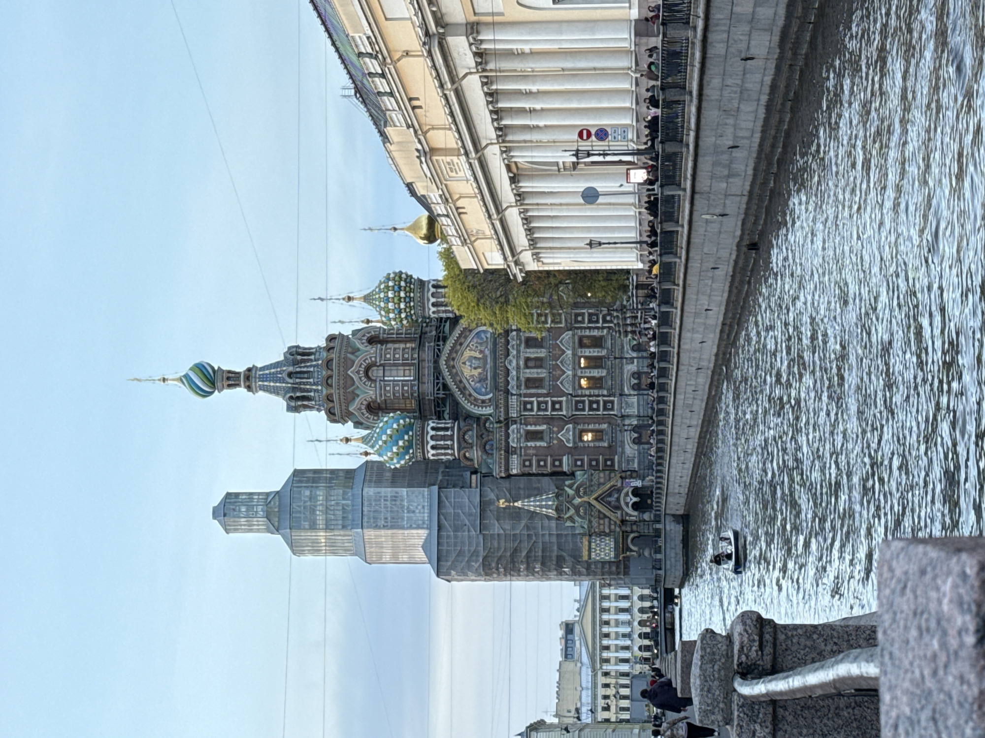 This image shows the Church of the Savior on Spilled Blood, located in Saint Petersburg, Russia. The church is partially covered in scaffolding. A river runs alongside the church, with a boat visible on the water. A building with columns is also beside the church. People can be seen walking along the embankment. The sky is light blue.