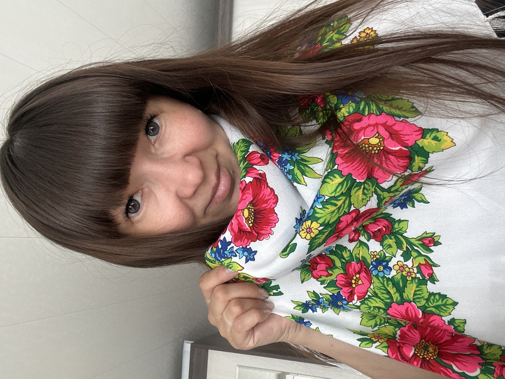 A woman with brown hair and a fringe is angled towards the camera. She is wearing a white shawl with a floral pattern including pink, red, blue and yellow flowers. The background appears to be a neutral-toned wall and a partial view of furniture. There's no explicit information tying the photo to the city of Среднее Кечево, Russia.