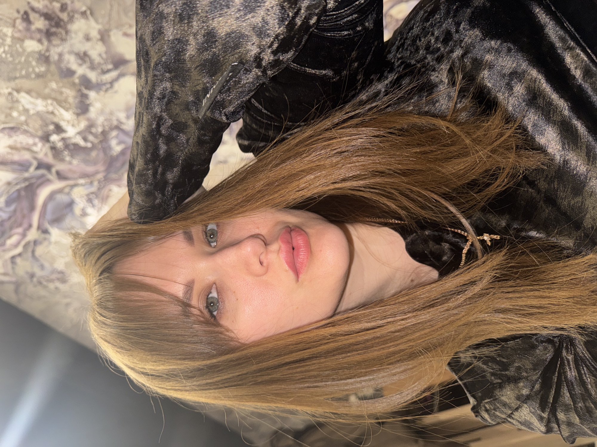 A portrait photo, presented rotated 90 degrees clockwise, features a young woman from the chest up. She has long, straight light brown hair with warm, reddish undertones and some lighter streaks, styled with soft bangs across her forehead. Her light-colored eyes (appearing grey or green) gaze directly at the viewer. Her fair complexion shows a few subtle freckles on her nose and cheek area. Her full lips are slightly parted and colored with a soft pink lipstick, giving her a pensive or alluring expression.
She is dressed in a dark, possibly black or very dark grey, crushed velvet garment that displays a textured, abstract pattern. A delicate gold-toned necklace with a small, star-shaped pendant is visible around her neck. On her clothing, near her shoulder, faint white text, possibly reading "INI", can be partially discerned.
The background is softly blurred, revealing a wall with an abstract or marbled pattern in tones of white, grey, and hints of lavender. The lighting is soft and diffused, indicating an indoor setting. The image captures a posed moment. This scene is located in Chaikovsky, Russia.