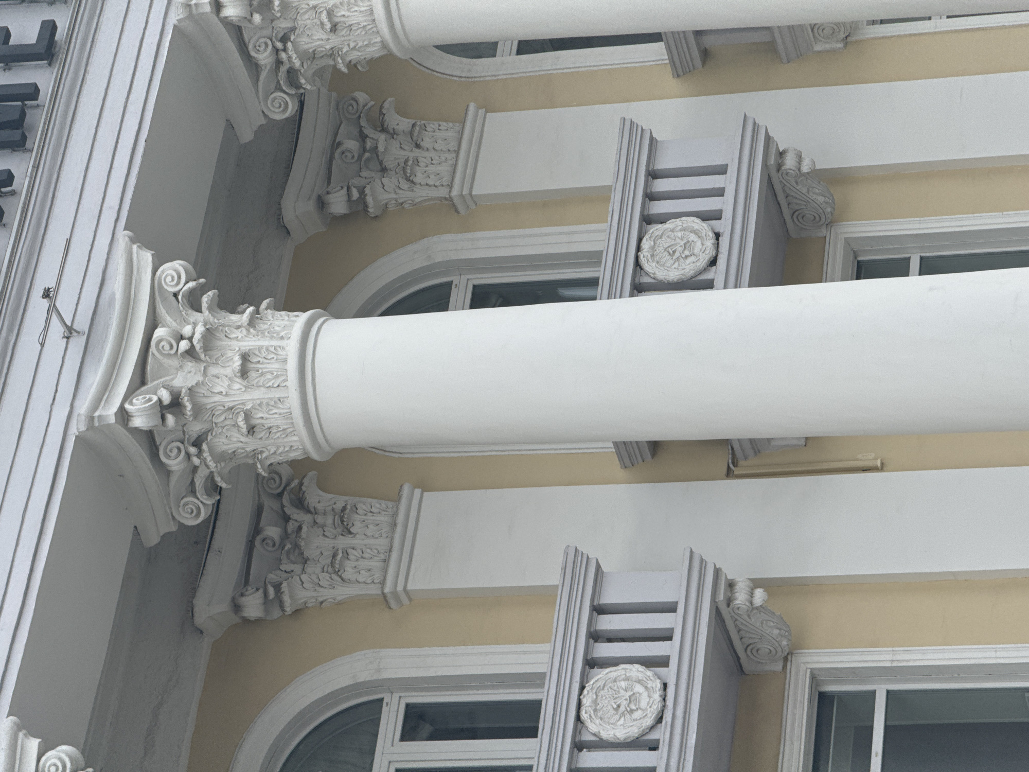 The image shows architectural details of a building in Krasnoyarsk, Russia. White, classical columns with ornate Corinthian capitals are visible. The building has cream-colored walls. Windows with arched tops and rectangular panes are present, along with decorative medallions and carved details.