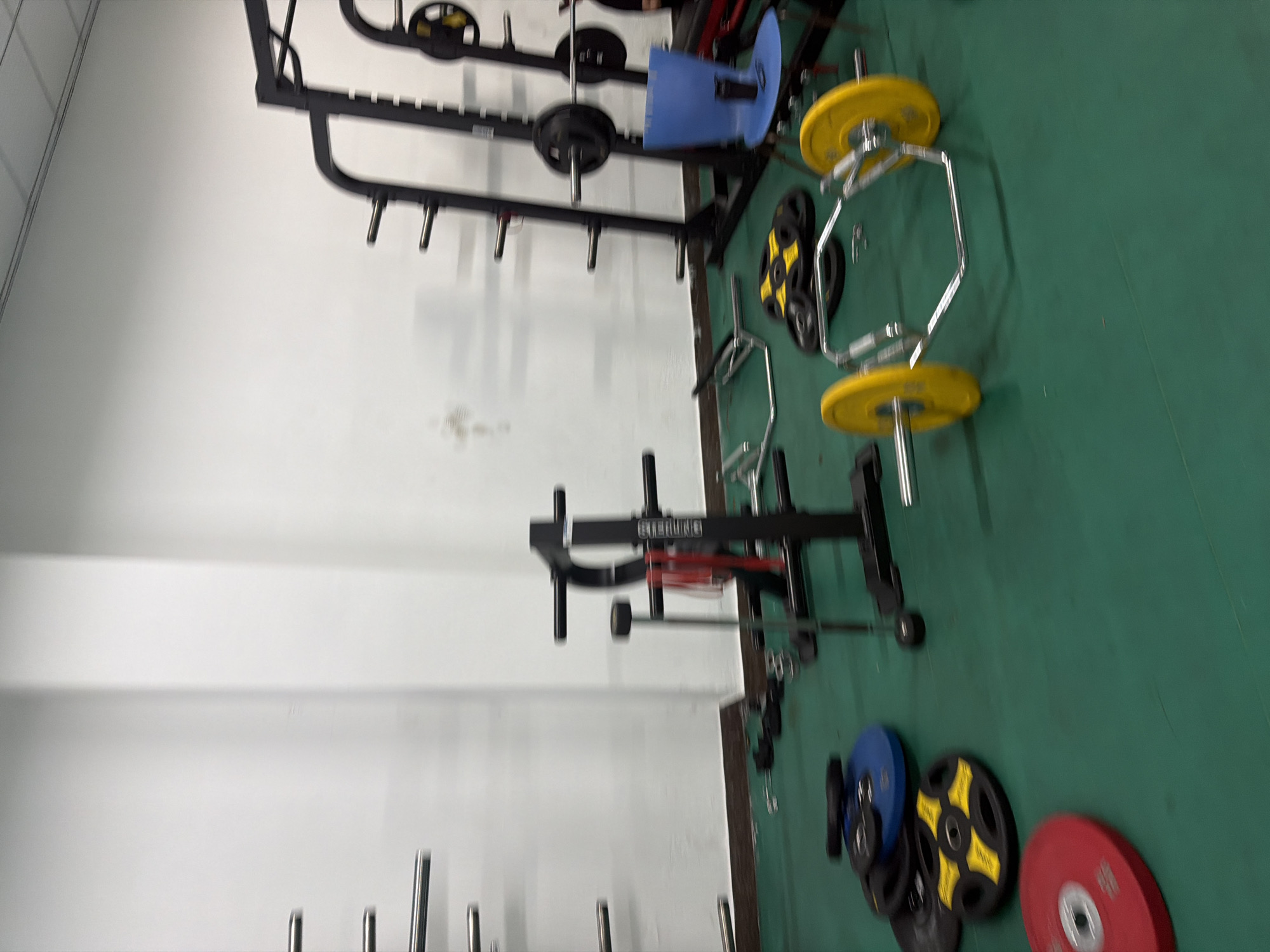 The image shows a gym with various weightlifting equipment. The equipment includes a power rack, a landmine attachment, a hex bar with yellow weight plates, and several weight plates of different colors (red, blue, yellow, and black). The gym is located in Jiangning District, China. The floor is green and the walls are white. There are no people visible in the image.