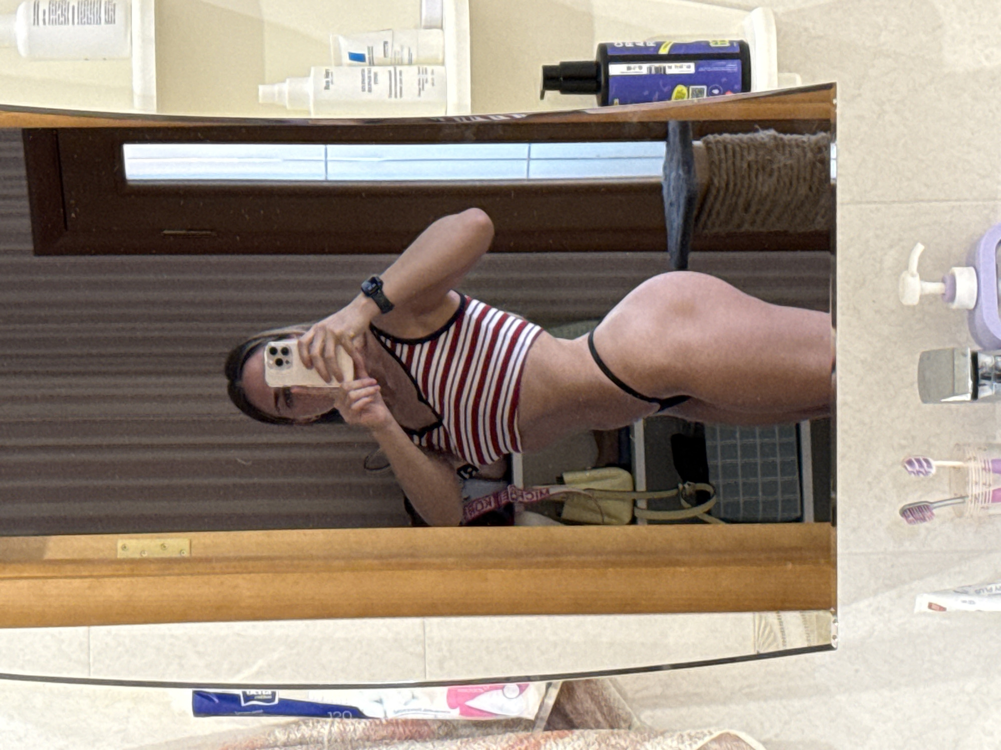 A reflection in a bathroom mirror shows a person taking a selfie. The person is wearing a striped top and black bikini bottoms. The bathroom contains toiletries on a shelf, a window with blinds, a cat tree, toothbrushes, and other items. The location is not discernible from the image, and there is no information to suggest it is in Saint Petersburg, Russia.