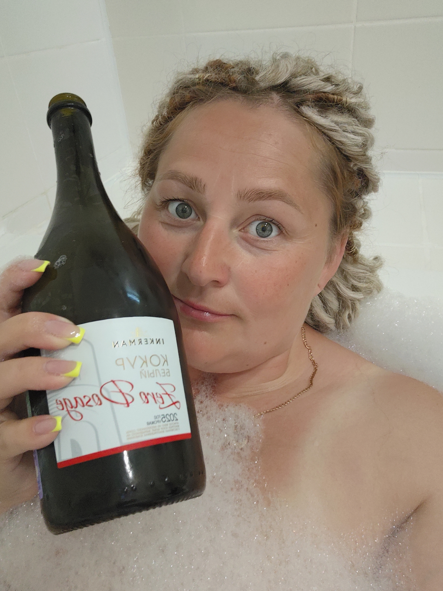A woman is in a bathtub filled with bubbles, holding a bottle of sparkling wine. She has curly blonde hair and is looking at the camera with wide, blue eyes. Her fingernails are painted neon yellow. The bottle has a label with text that appears to be in Russian and English, and some of the English text is upside down. The environment is a bathroom with white tiled walls. The image was taken in Sochi, Russia.